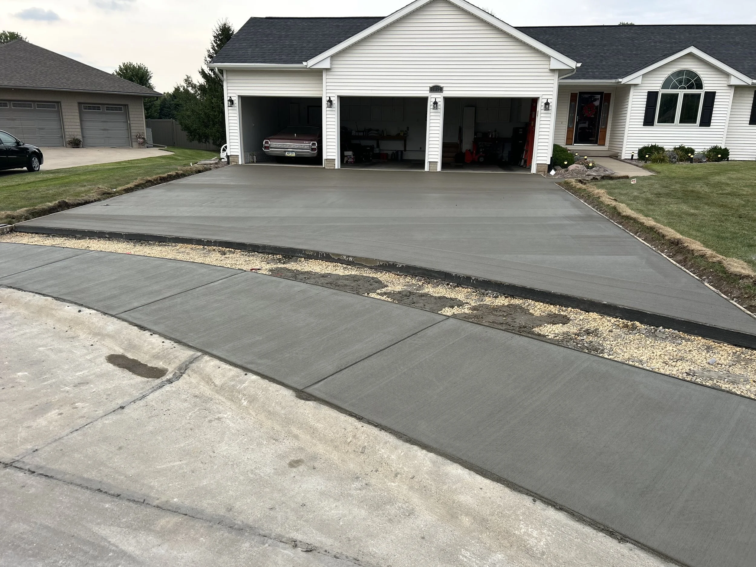 This image shows a residential driveway with a newly poured concrete section. The house has a white exterior with black shutters and a front door decorated with a wreath. The driveway is in the process of being finished, with some areas still needing
