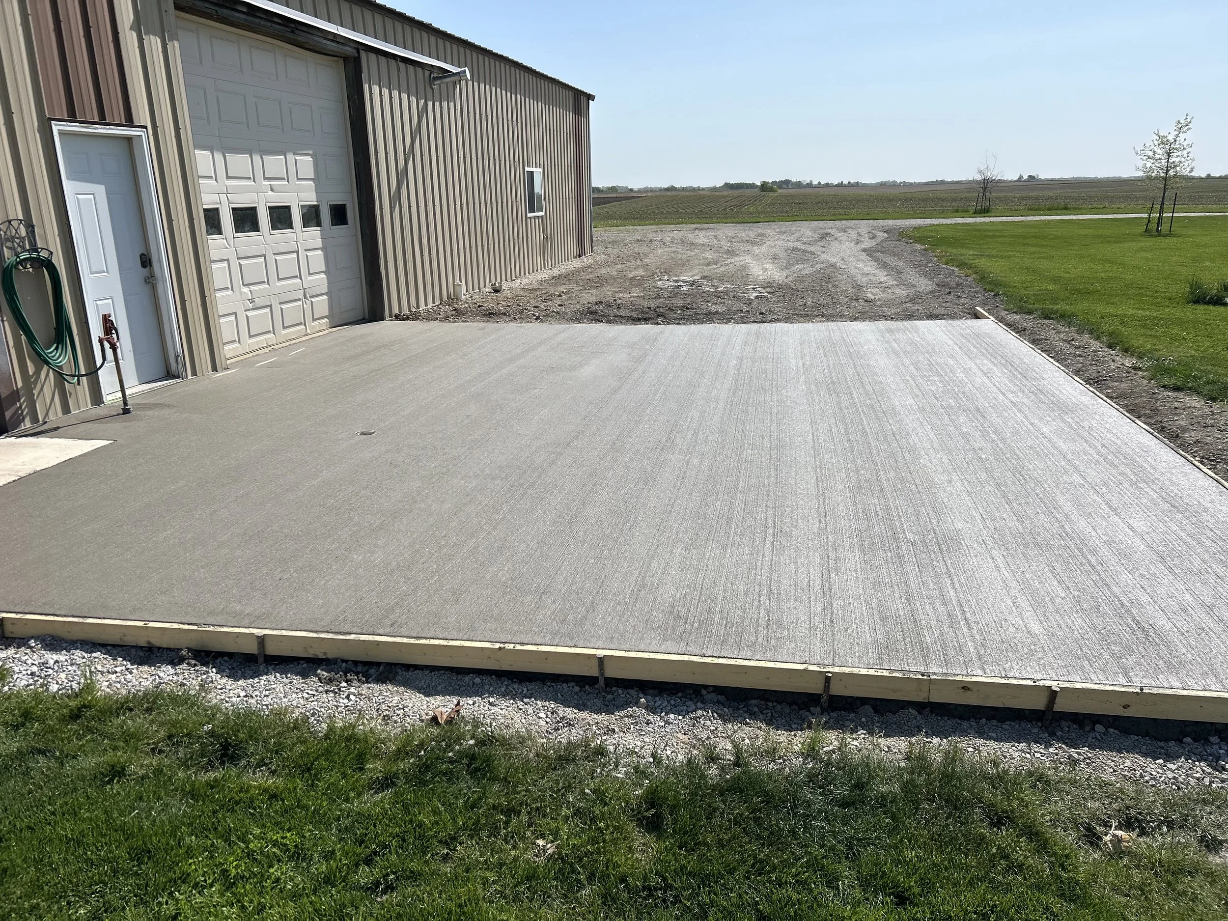 A partially constructed concrete porch in front of a metal building with a large garage door and a small side door, situated on a rural property with open fields and a gravel driveway.