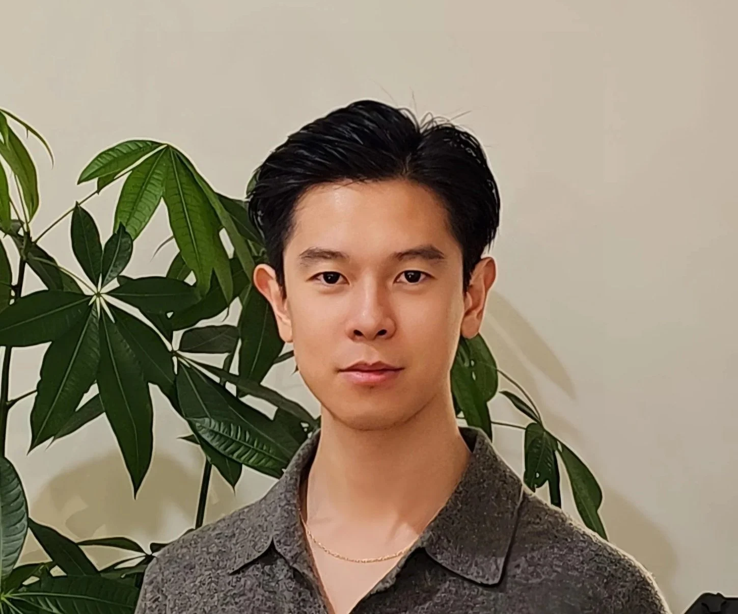A young man with black hair and light skin standing in front of green leafy plants against a plain beige wall.