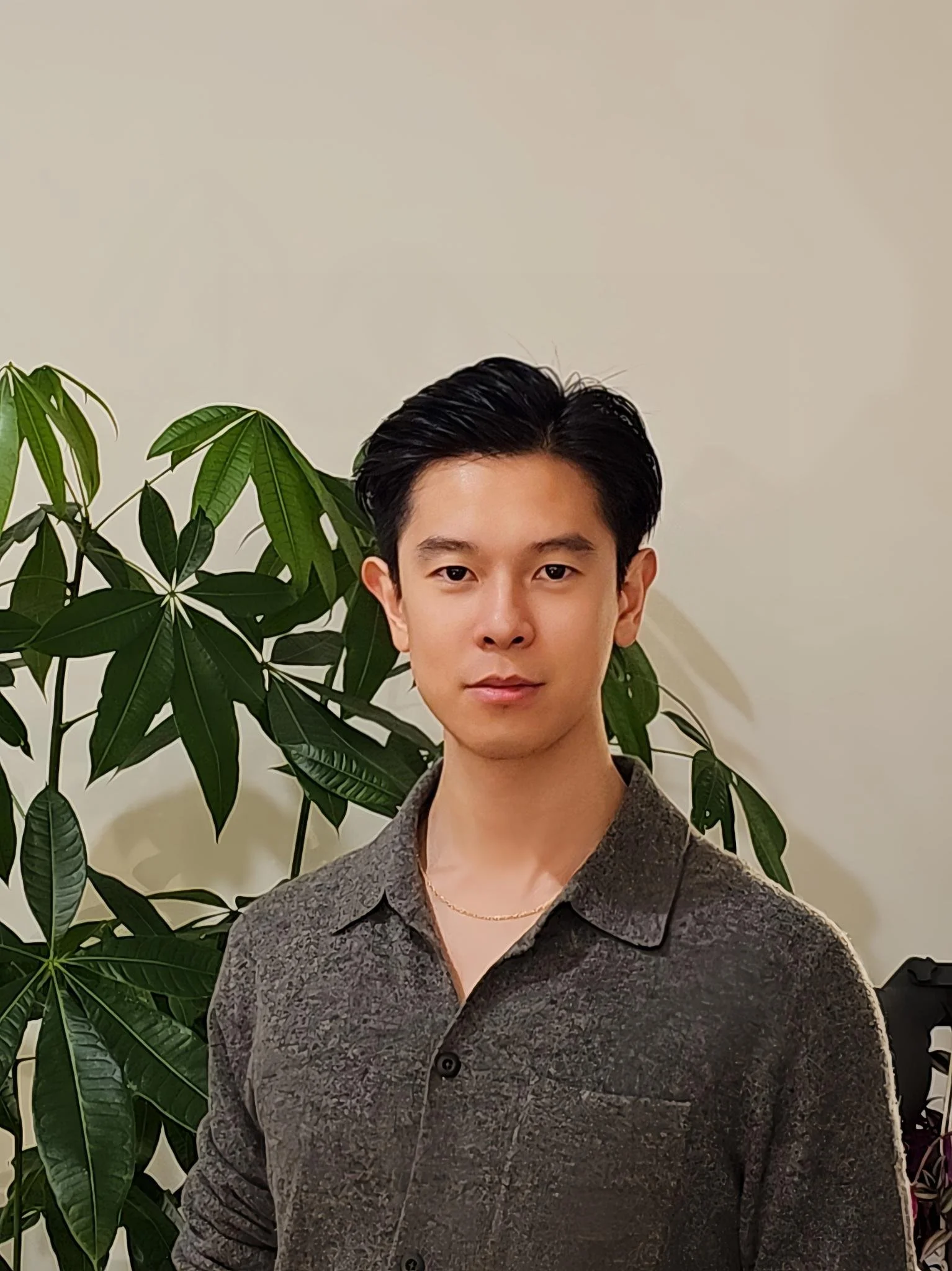 A young man with black hair and a focused expression standing in front of a large green plant with palm-like leaves and a plain light-colored wall.