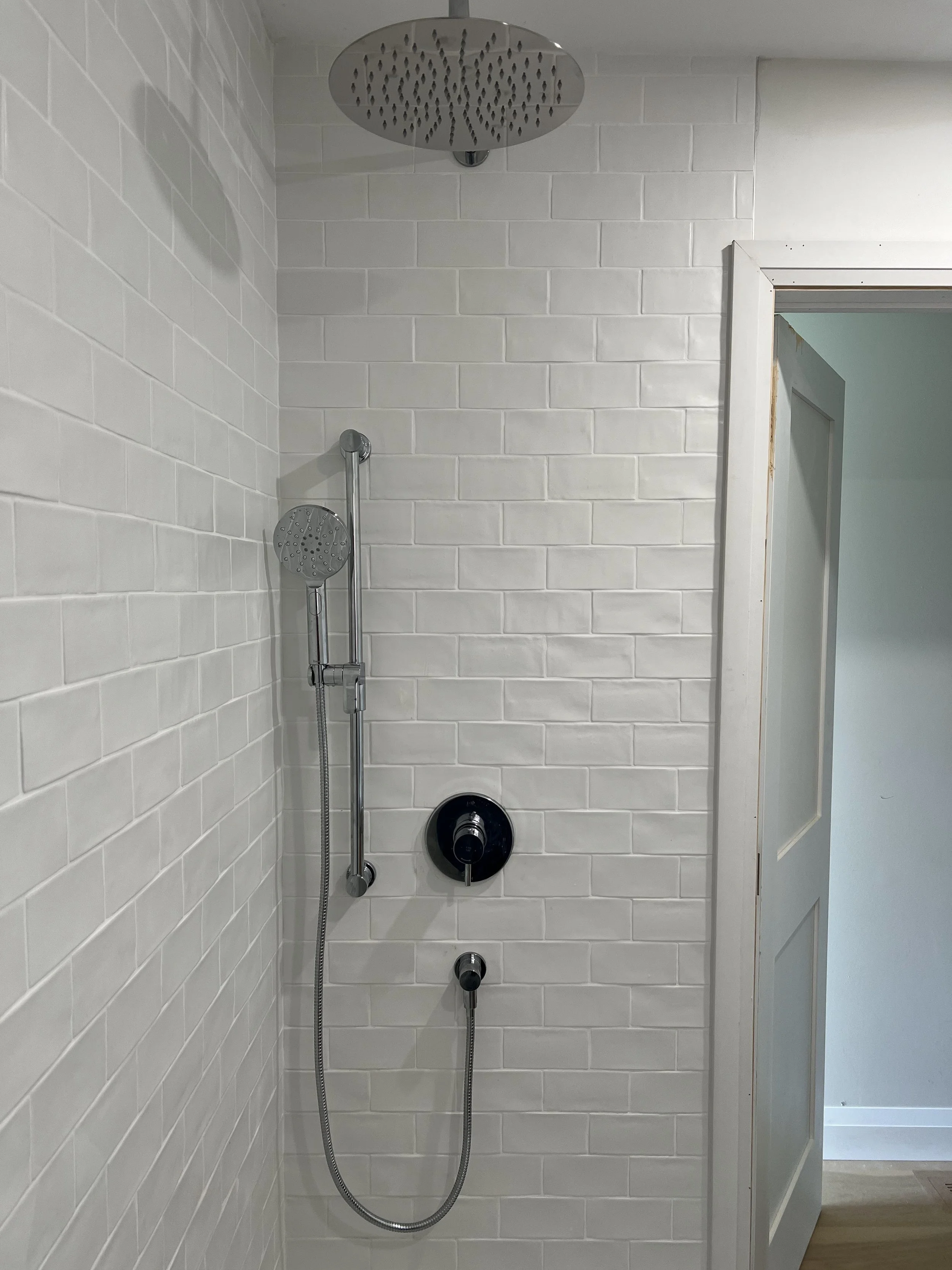 Shower area with white tiled walls, a modern rain shower head mounted at the ceiling, and a handheld shower head attached to a sliding bar on the wall. A door is partially visible to the right.