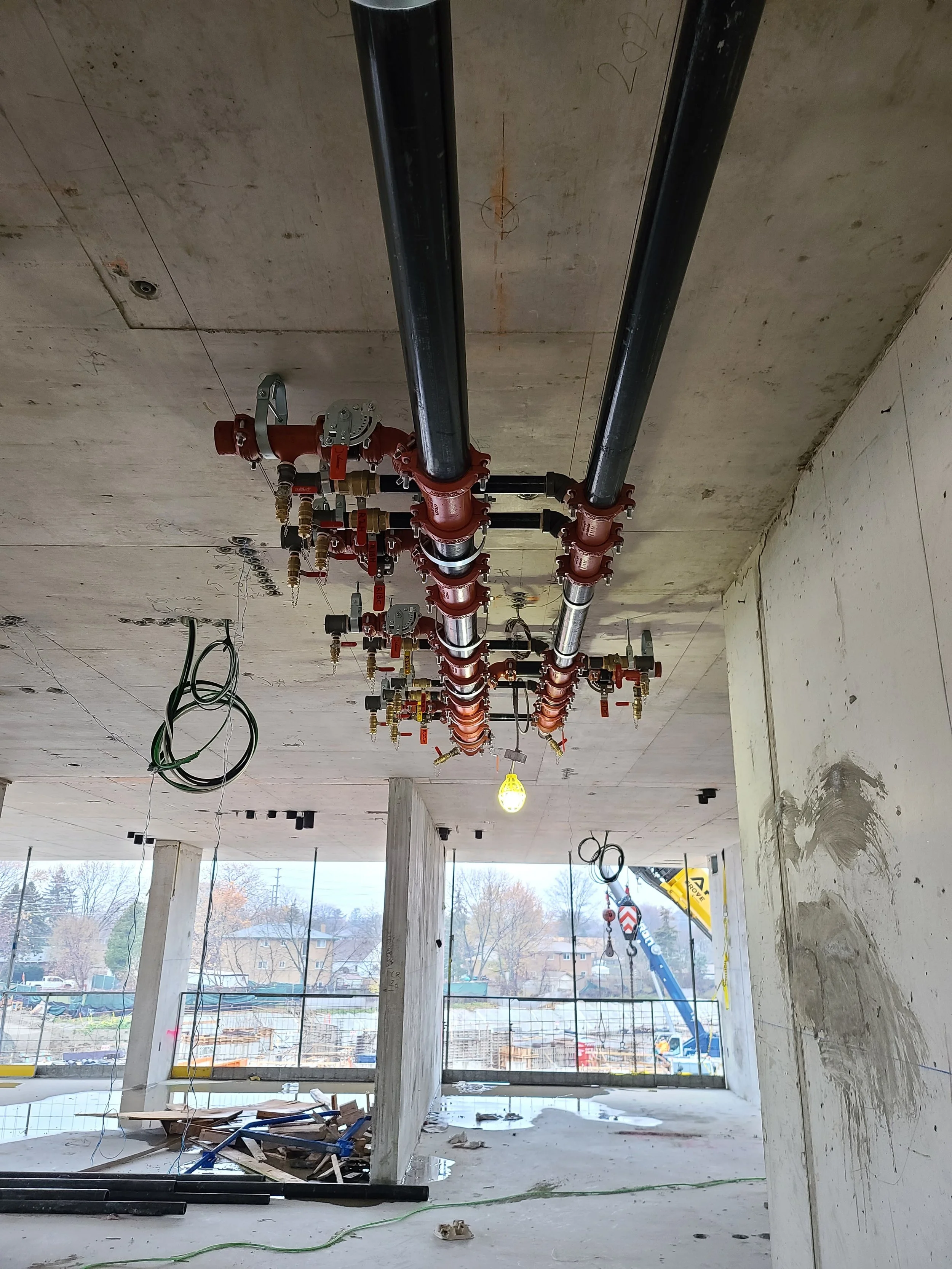 Construction site with exposed pipes and wiring inside a building under construction. Large windows reveal a view of trees and buildings outside.