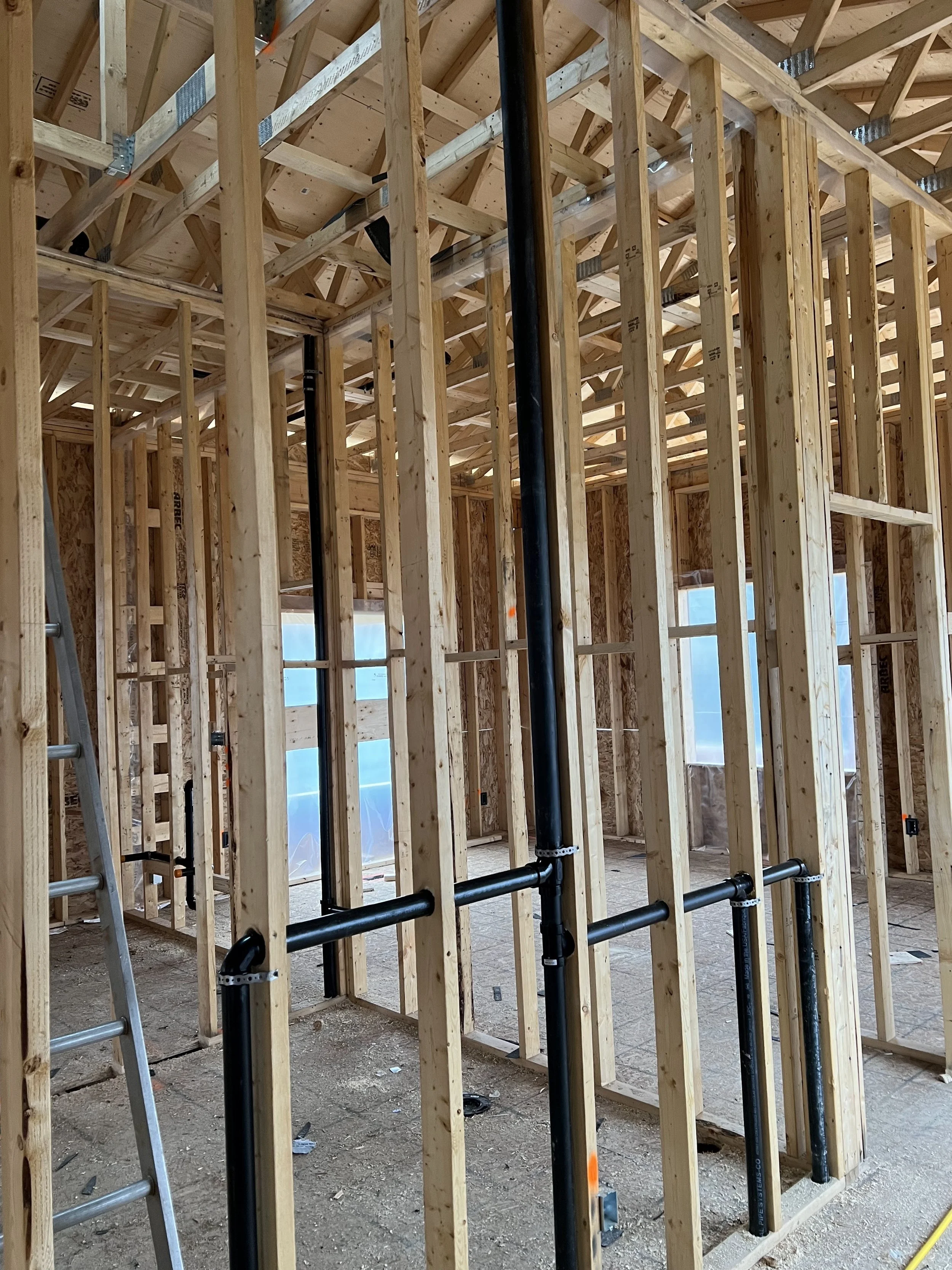 Interior view of a building under construction with wooden framing, black plumbing pipes, and a ladder.