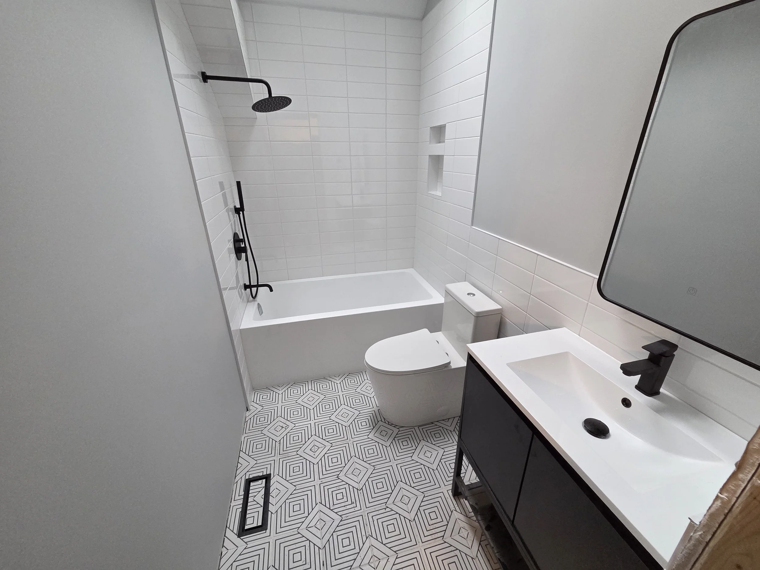 Modern bathroom with a shower, toilet, and vanity sink. The shower has a black rain showerhead, a handheld shower, and built-in niches for storage. The floor has a geometric black and white tile pattern, and the walls have white rectangular tiles.