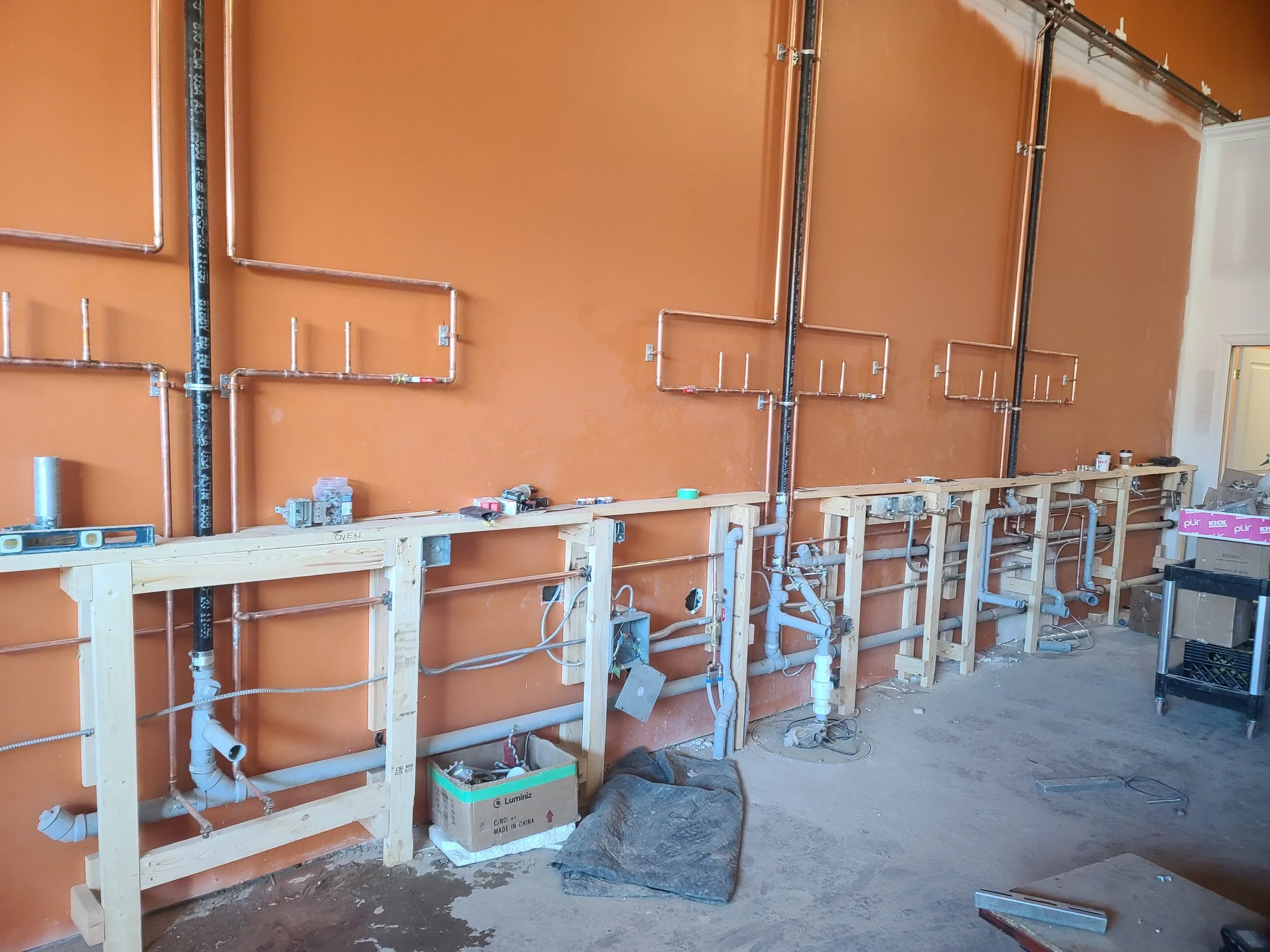Construction site with exposed plumbing pipes and wooden framing against an orange wall.