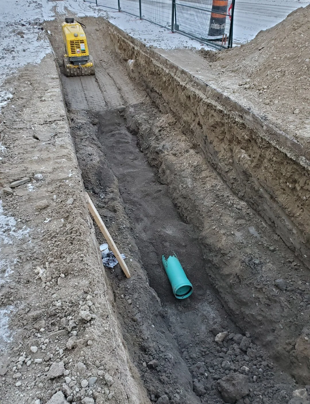 Construction site with a deep trench, a small yellow plate compactor, and a green pipe in the trench.