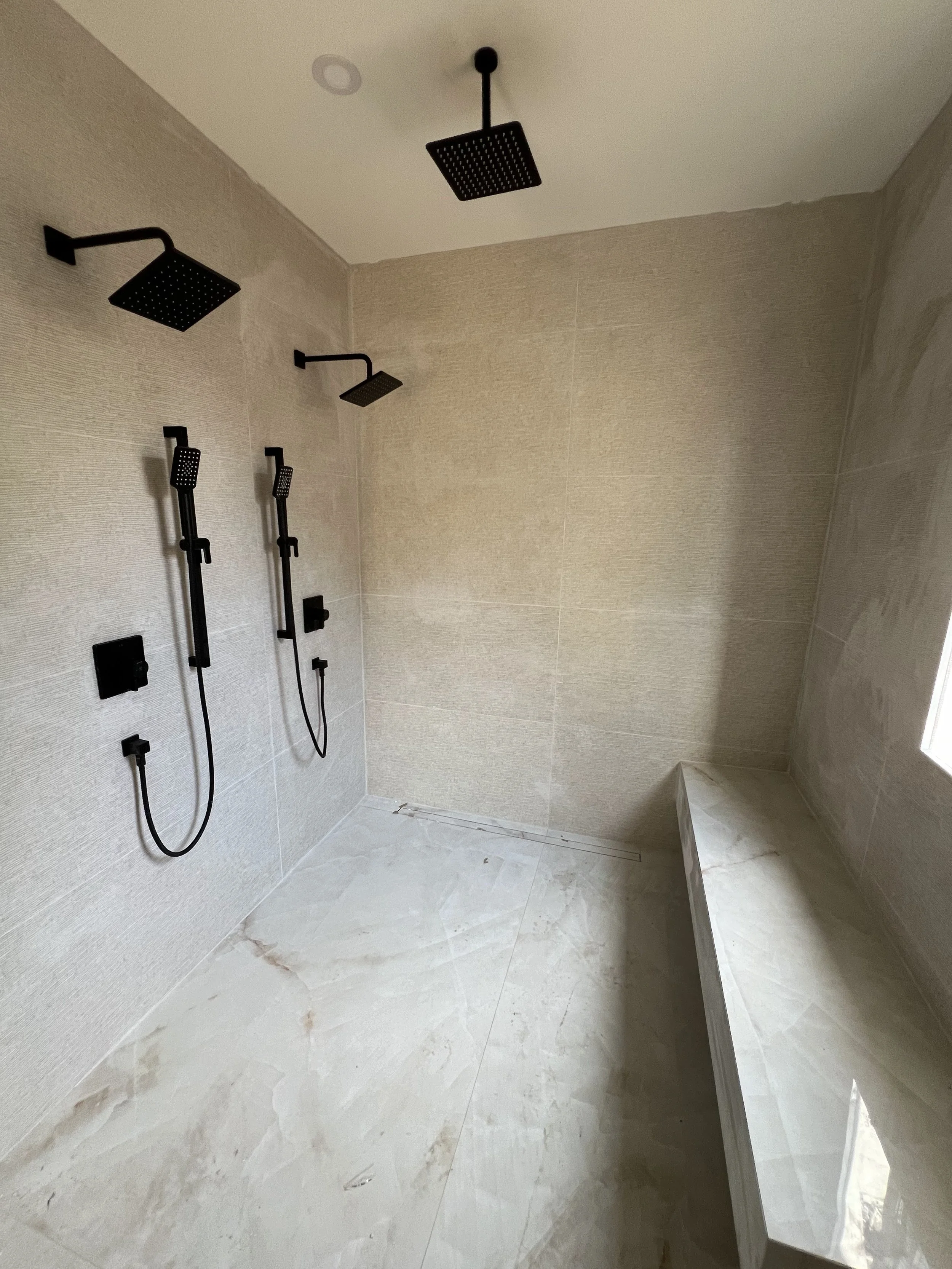 A modern shower area with three black rain showerheads mounted on the beige wall, two black handheld showerheads on the left, and a marble bench on the right side, with beige tiled walls and floor.