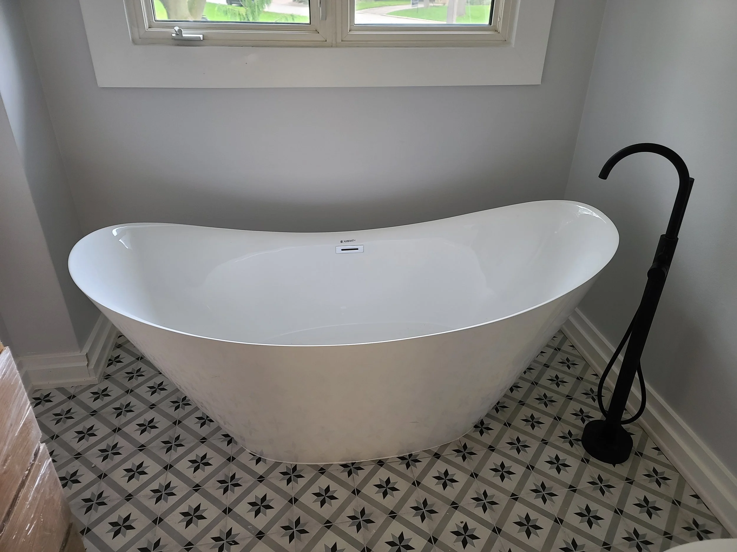 Modern white freestanding bathtub with black faucet near window in a bathroom with patterned tile floor.