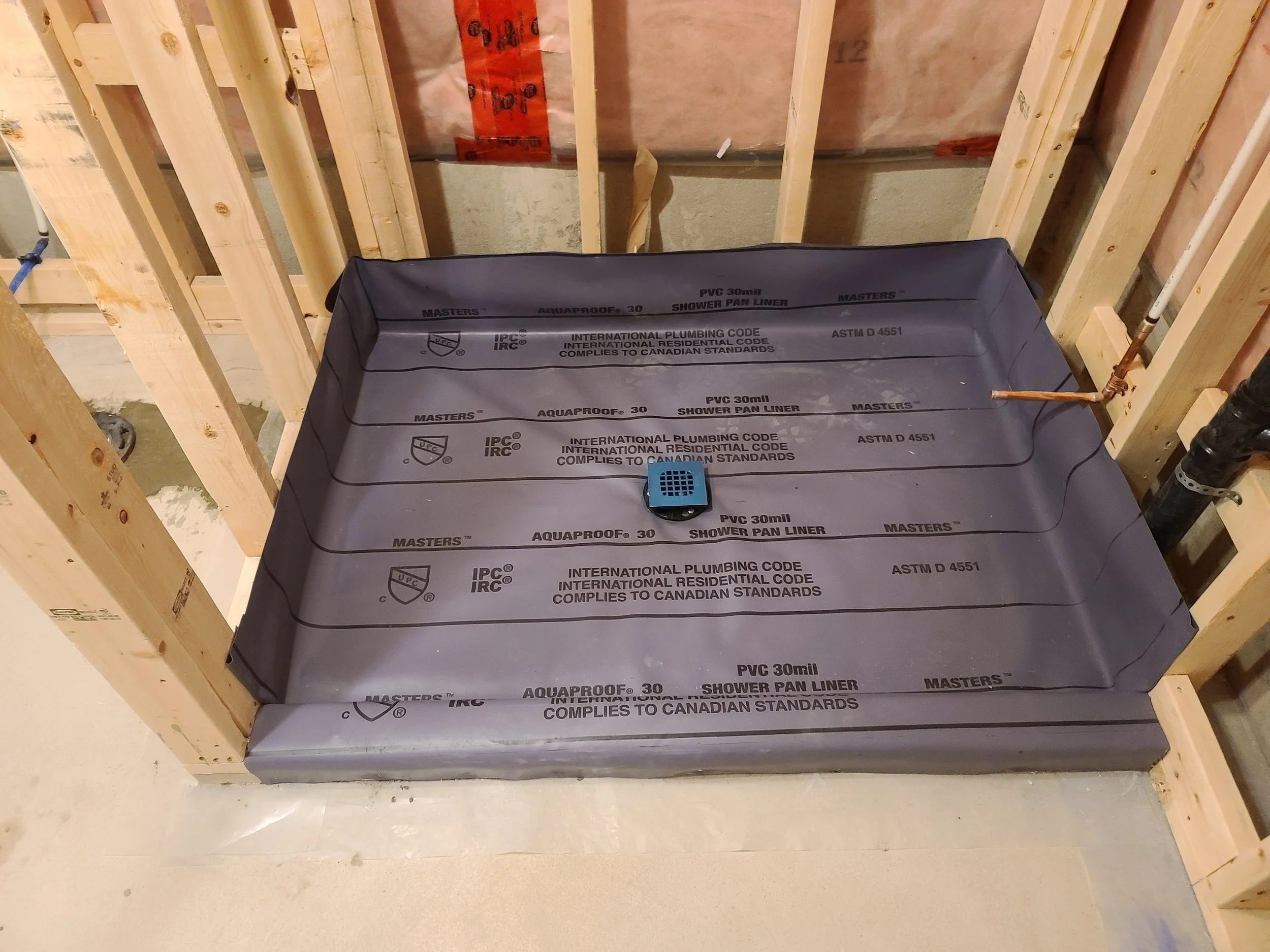 Construction framing with wooden studs and a black shower pan liner labeled 'Masters Aquaproof 30' placed in the bathroom floor.