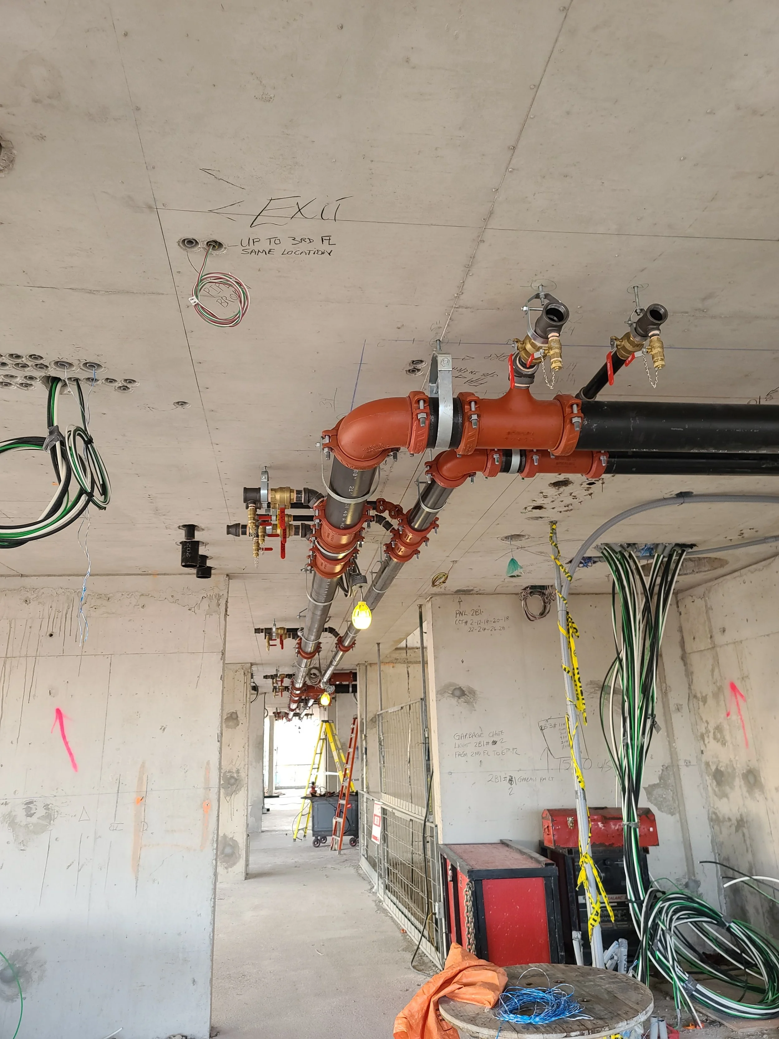 Construction site with exposed pipes, wires, and electrical wiring on a concrete ceiling and walls.