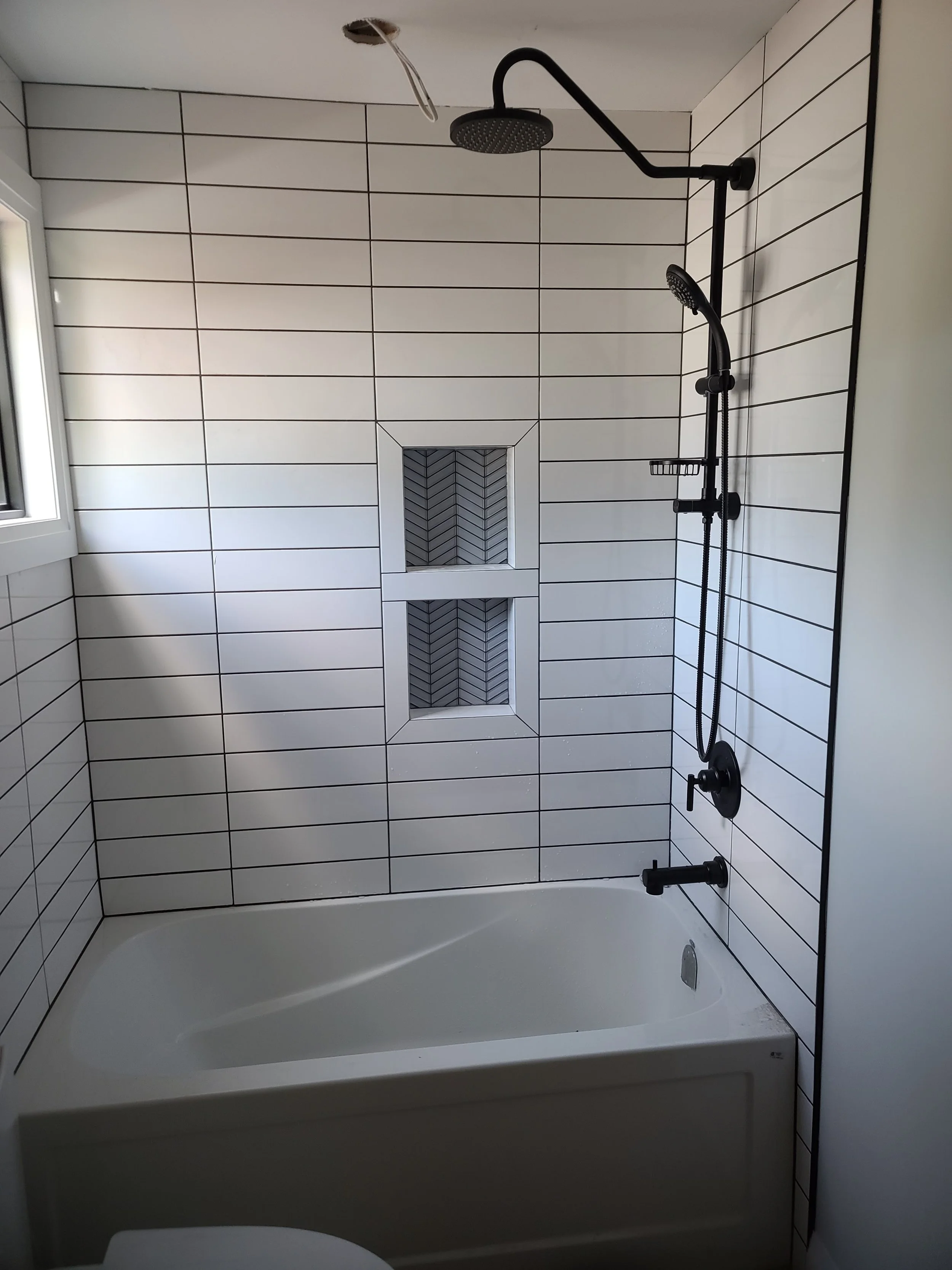 Bathroom with bathtub, black shower fixtures, and white tiles with black grout, including a small built-in shelf with a herringbone pattern.
