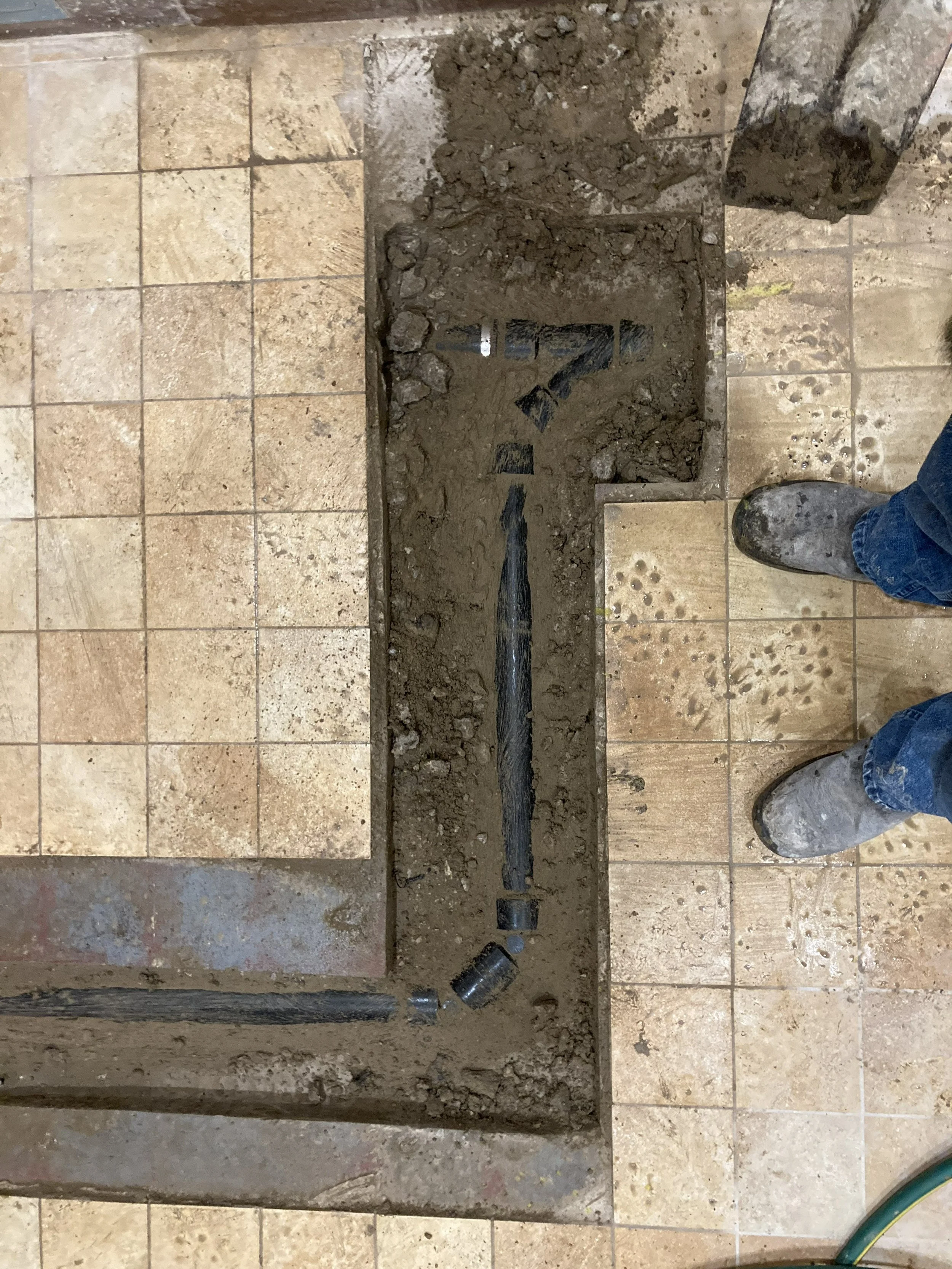A floor with removed tiles revealing the subfloor, dirt, and a pipe, indicating an ongoing renovation or repair.