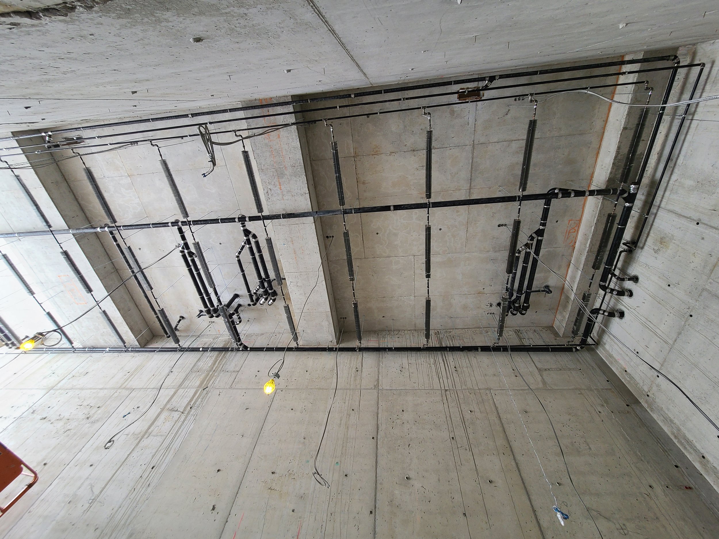 Construction site ceiling with exposed black pipes, electrical wiring, and construction lights, unfinished concrete walls, and visible ceiling fixtures.