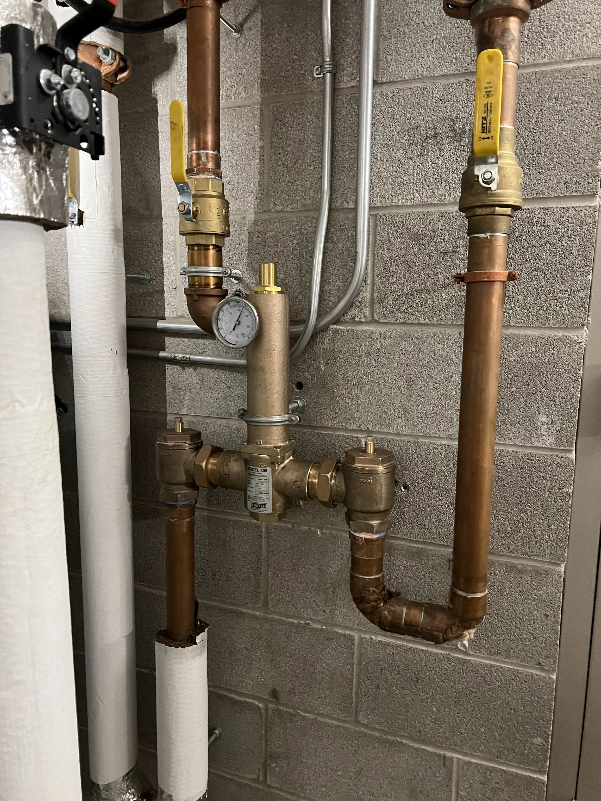 A plumbing system with copper and PVC pipes, a pressure gauge, and yellow-handled shut-off valves mounted on a concrete block wall.