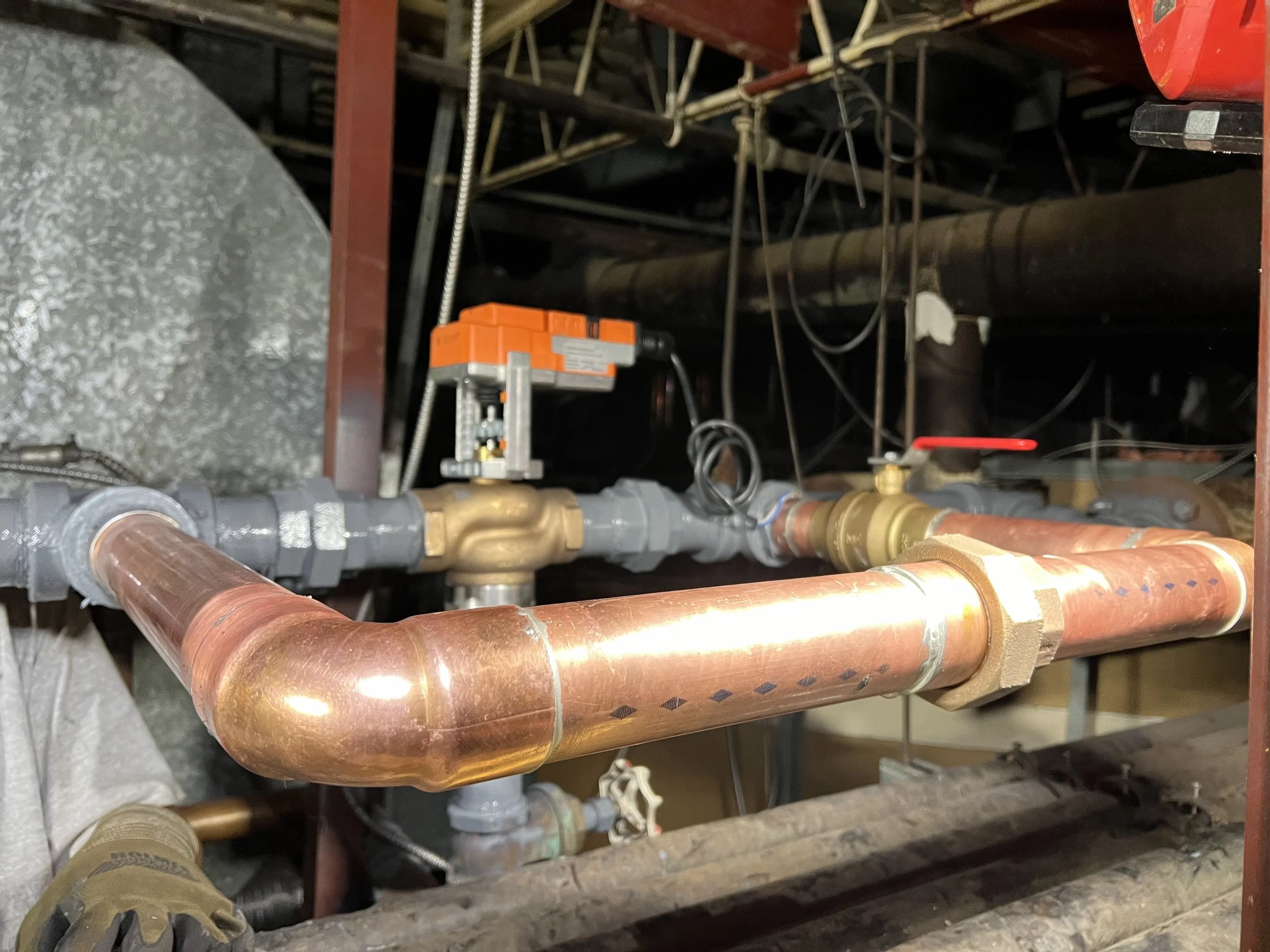 A network of copper pipes with valves, gauges, and wiring in a mechanical or plumbing system, with some insulation on one pipe and a person's foot visible at the bottom.