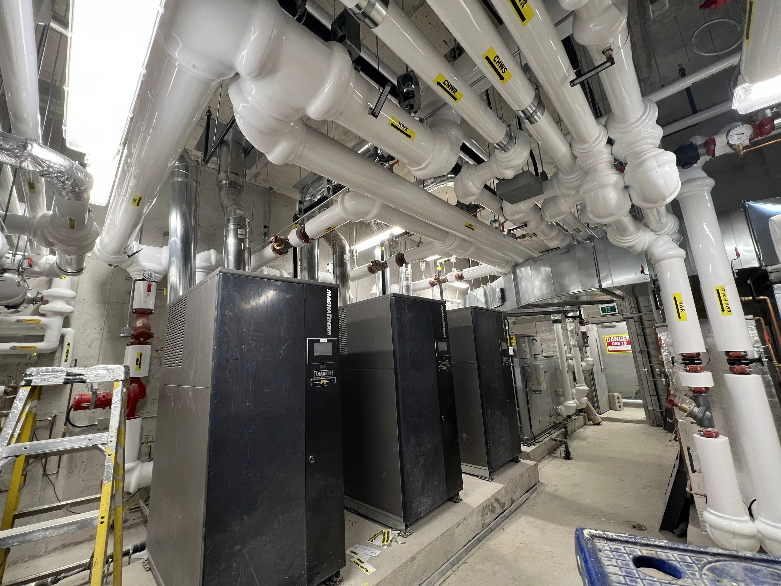 A mechanical room with large white pipes, valves, and industrial equipment, including a black control box, yellow warning tags, and a ladder.