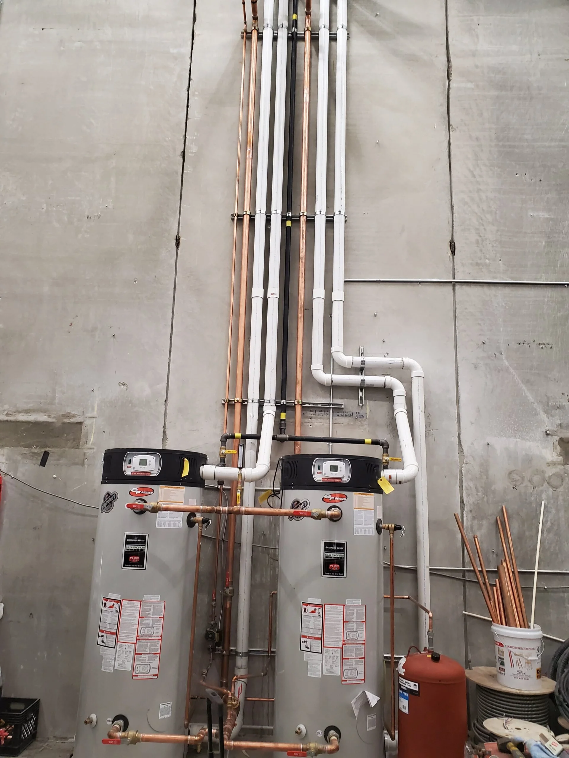 Two water heaters with various copper and white PVC pipes connected, set against a concrete wall in a utility room.