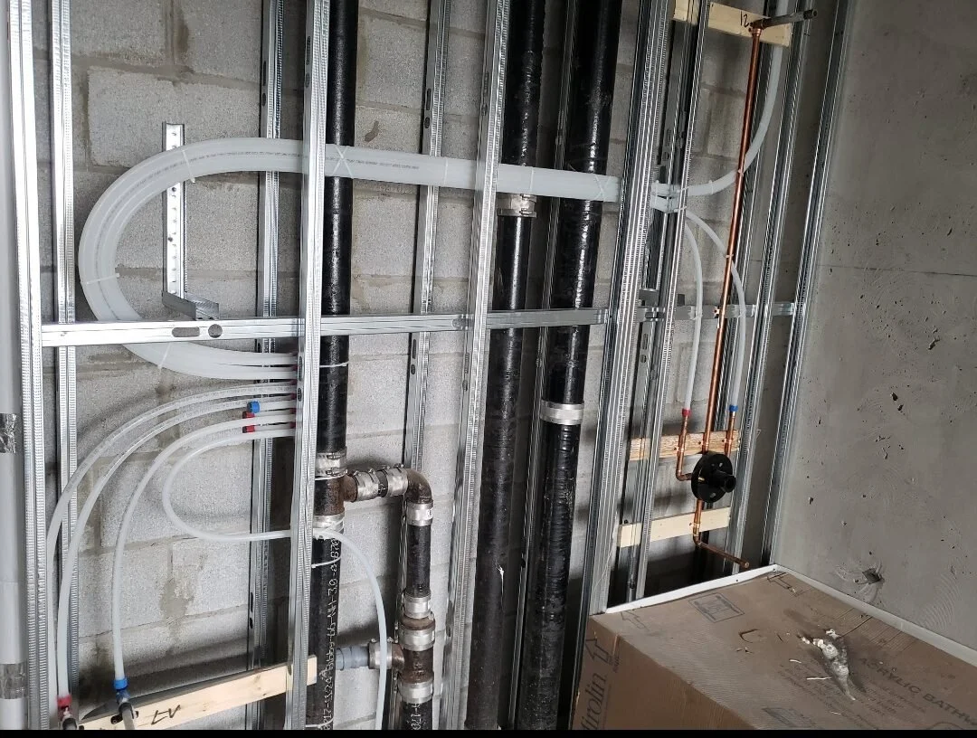 Construction site with metal framing, black and copper pipes, and plastic tubing installed on a wall.