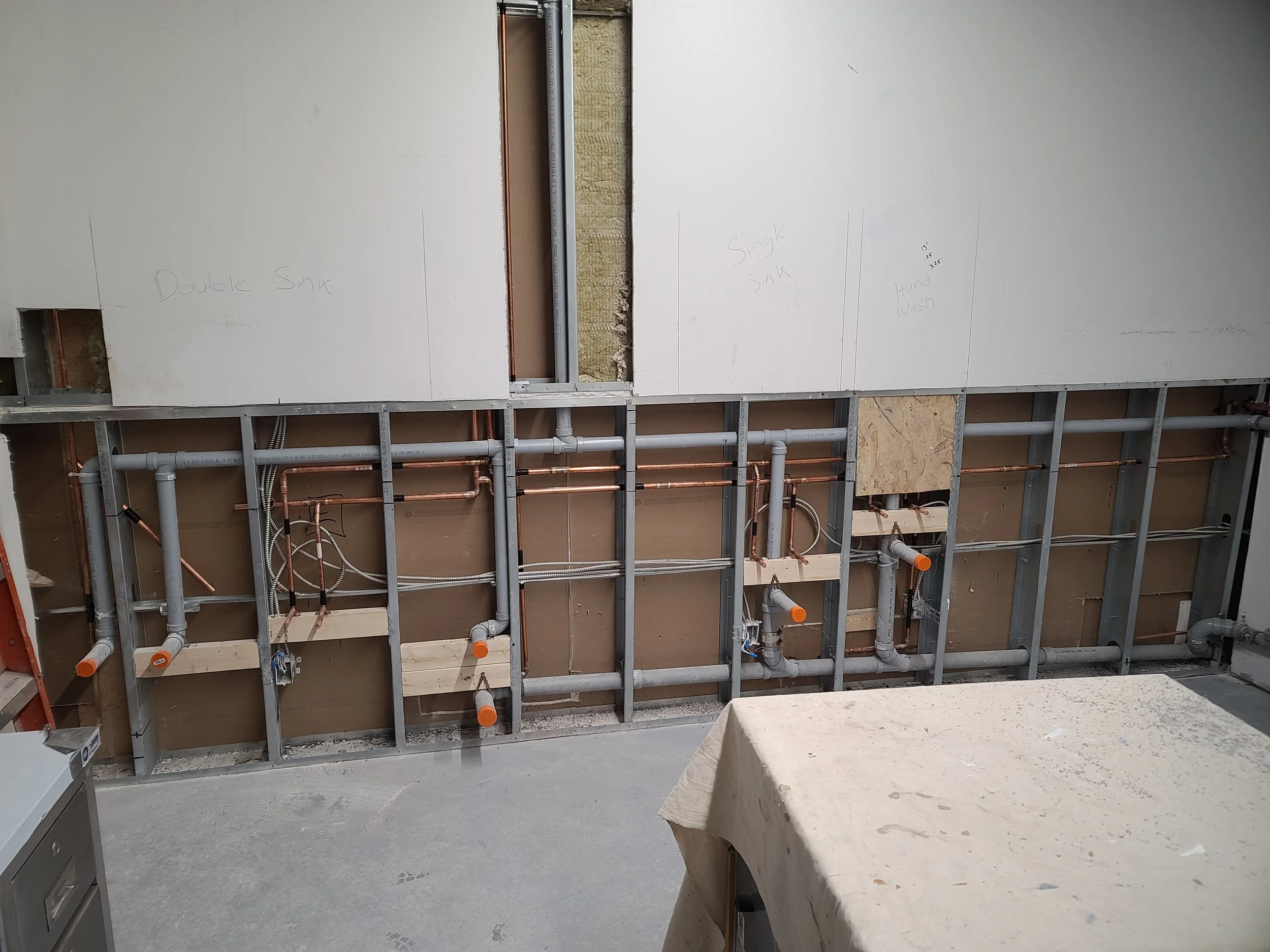 Construction site showing interior wall framing and plumbing pipes, with some drywall panels marked with handwritten labels like "Double Sink," "Single Sink," and "Hand Wash."