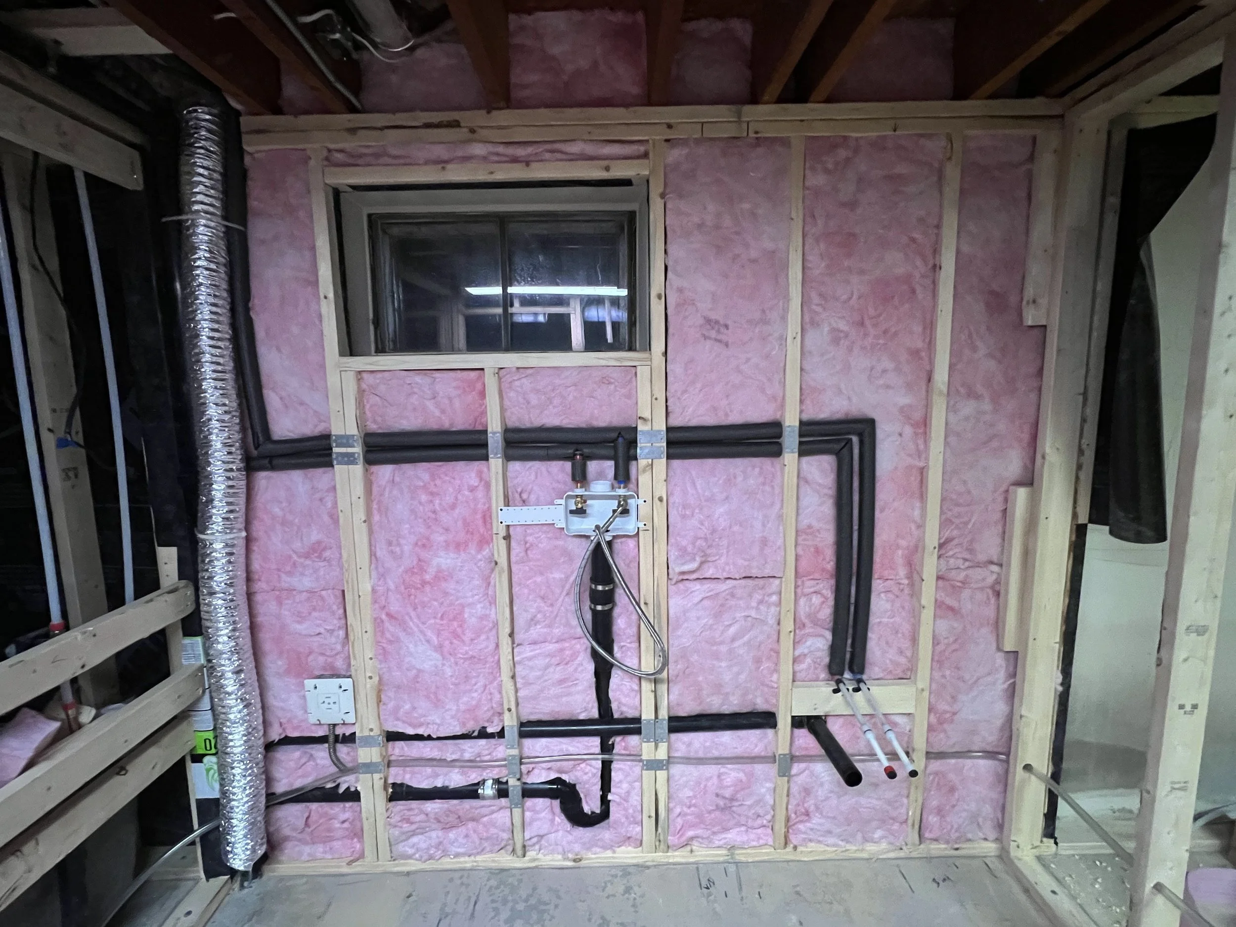 Interior of a room under construction with pink insulation, wooden studs, and various pipes and fixtures installed.