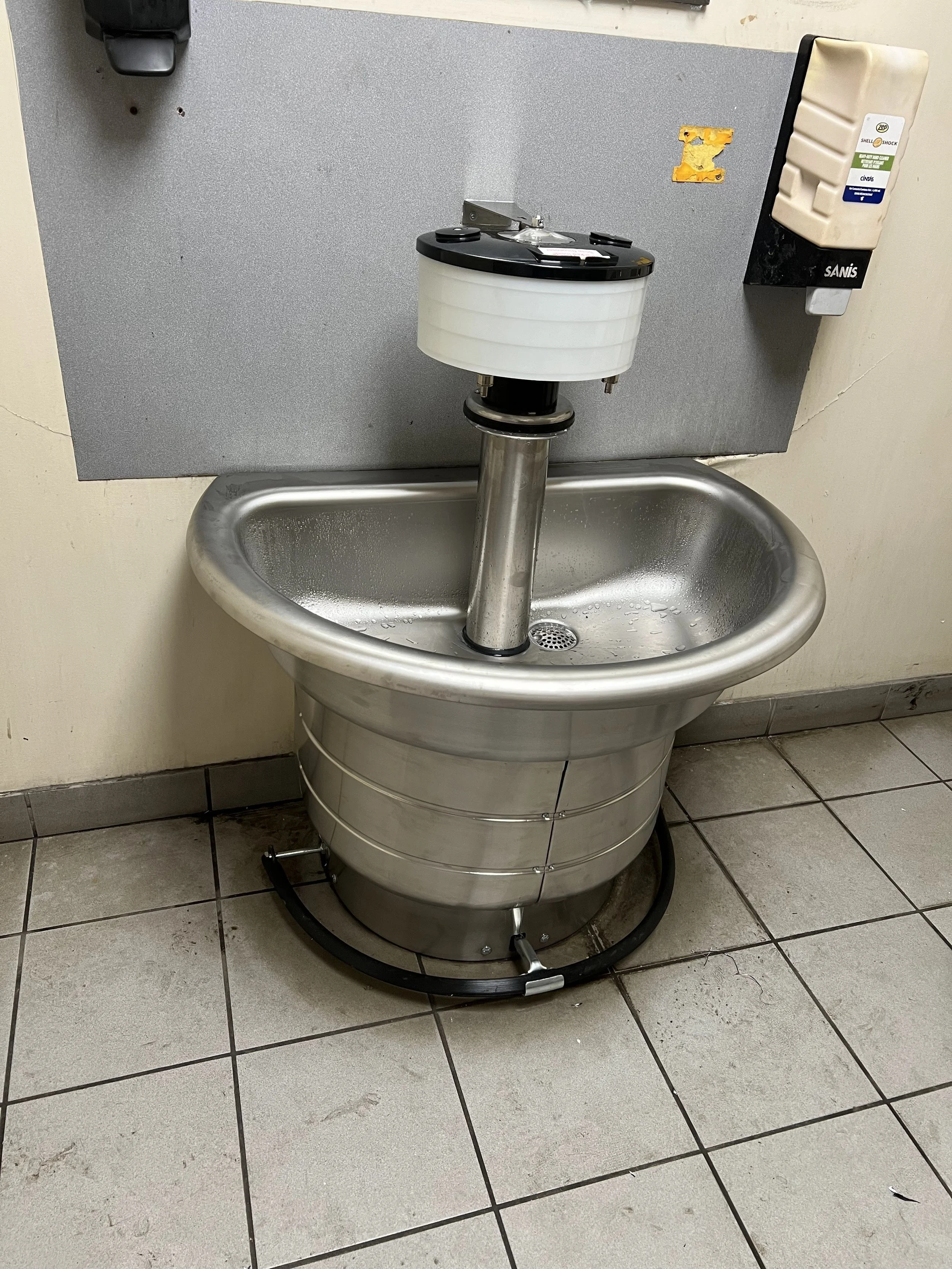 A stainless steel medical sink with a faucet and soap dispenser mounted on a wall with tile flooring.