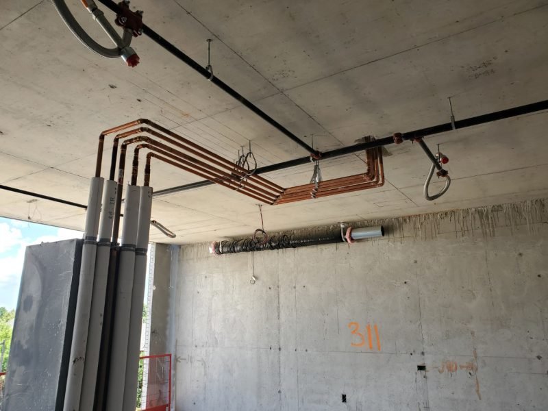 Construction site ceiling with exposed copper pipes, electrical conduits, and a ventilation duct, with a concrete wall in the background.