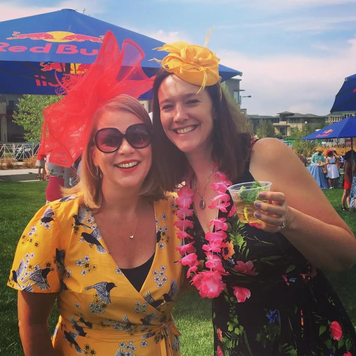 Two women dressed in colorful, floral, and festive outfits, standing outdoors at a party or celebration with umbrellas and other people in the background.