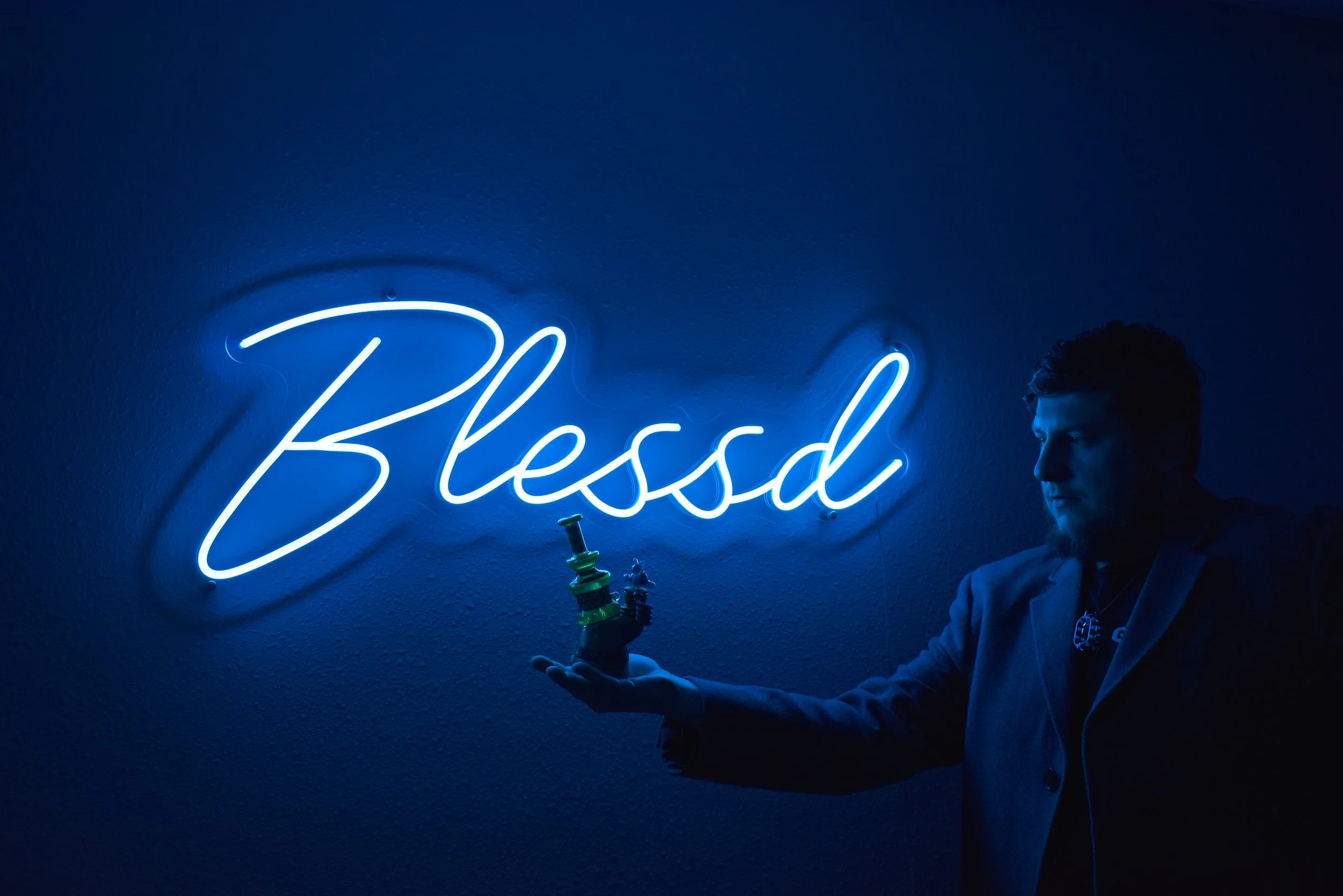 A person with dark hair and a beard wearing a light-colored blazer is holding a science apparatus with a yellow and green glass container. Behind him, there is a glowing neon sign on the wall reading 'Bless' in cursive blue letters.