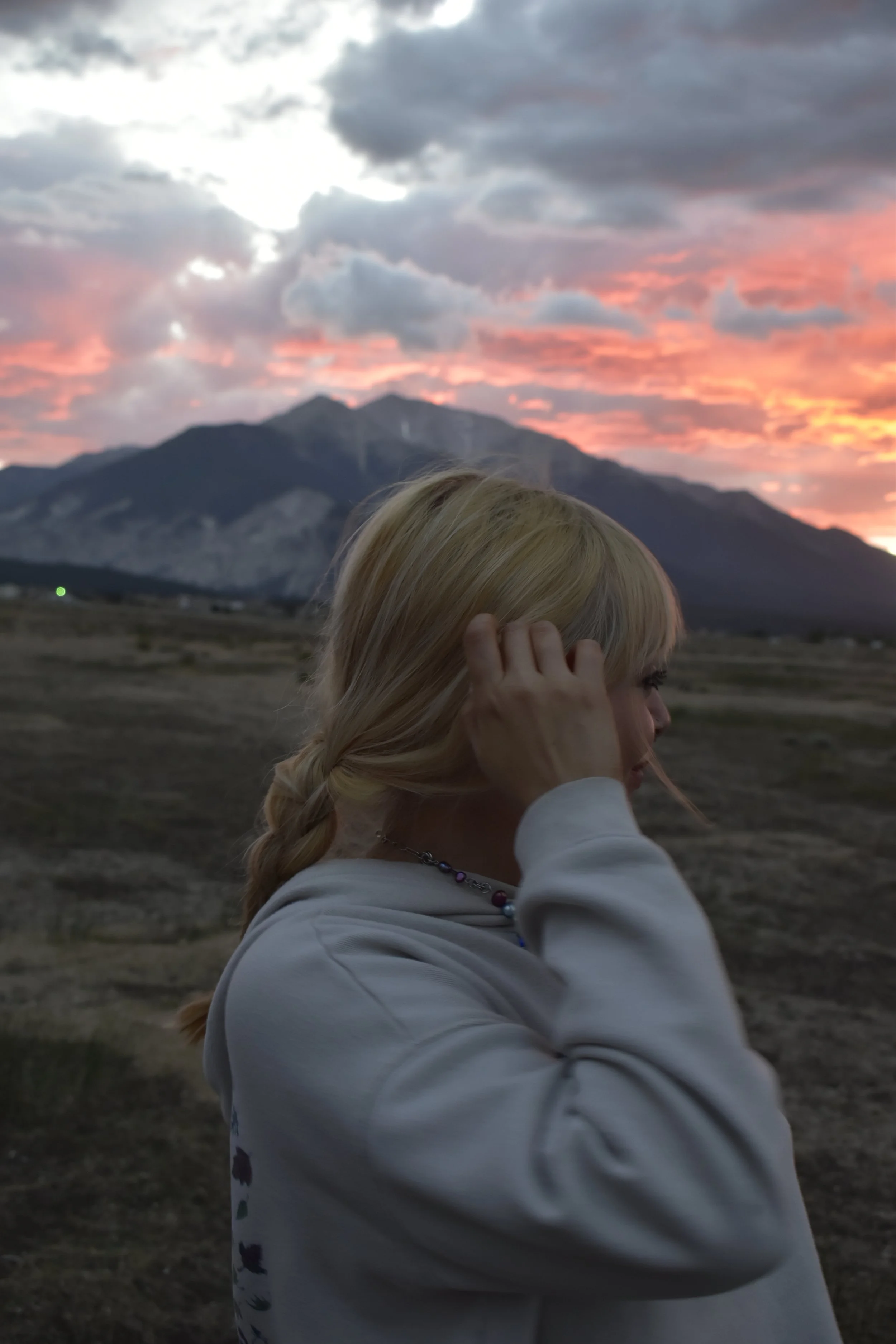 A woman with blonde hair styled in a braid is touching her head, standing outdoors on a land with mountains in the background during sunset, with a sky filled with colorful clouds.