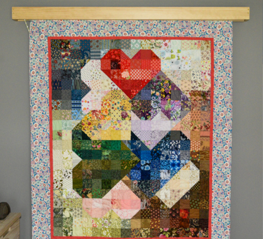 Colorful quilt hanging on a rod with floral and geometric patterns, framed with a red border, against a gray wall.