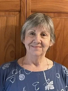 Smiling elderly woman with short gray hair wearing a blue top, standing in front of wooden cabinets.