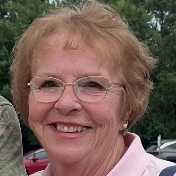 A smiling elderly woman with short, wavy hair and glasses, wearing a dark jacket and a white shirt, sitting outdoors at a restaurant or cafe near a parking lot with cars and an ethanol fuel sign in the background.