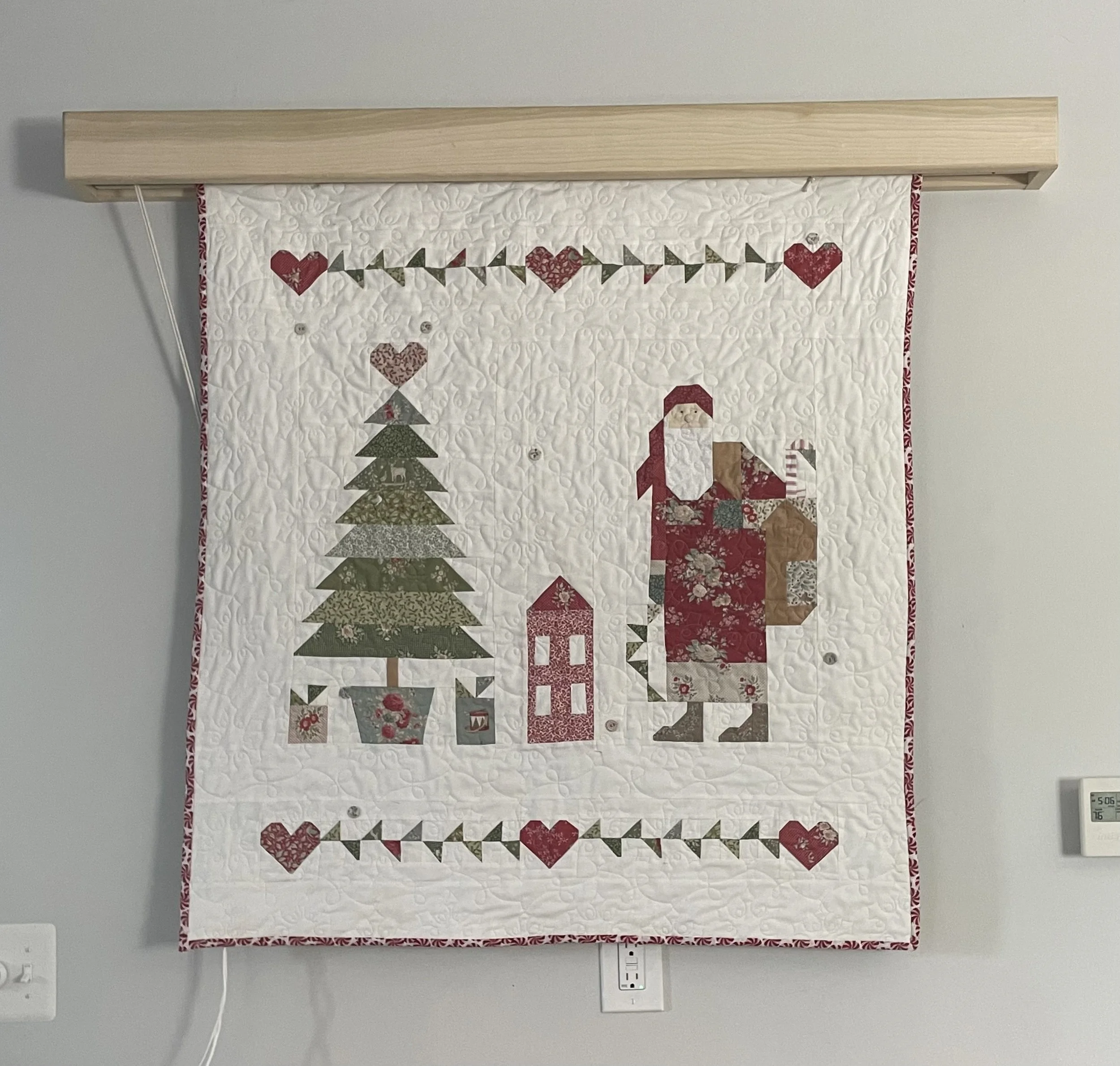 A quilted wall hanging with Christmas-themed patterns, including a Christmas tree, Santa Claus, a gift box, and hearts, bordered with red and white fabric, hanging on a wall with a light gray background. The quilt is attached to a wooden dowel.