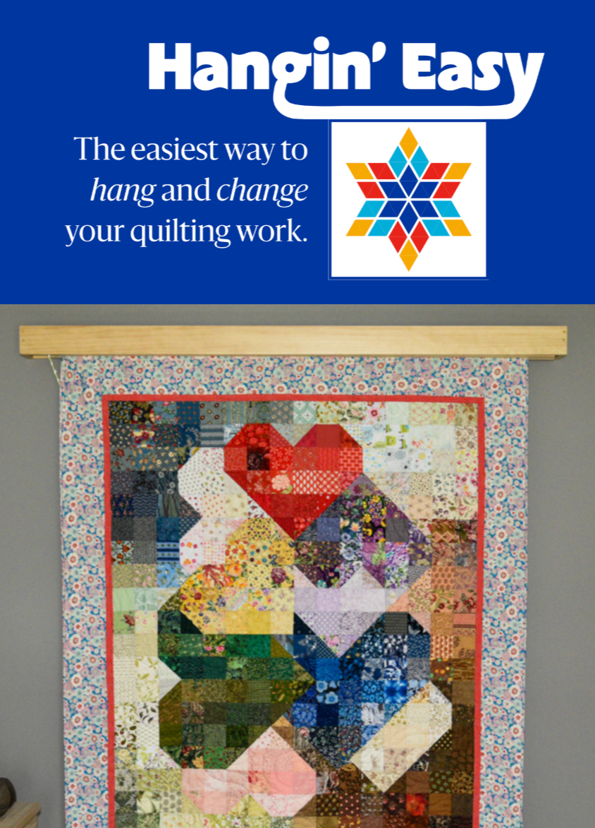 A colorful quilt hanging on a wooden rod with a blue sign above it that says 'Hangin’ Easy' and explains that it is an easy way to hang and change quilting work.