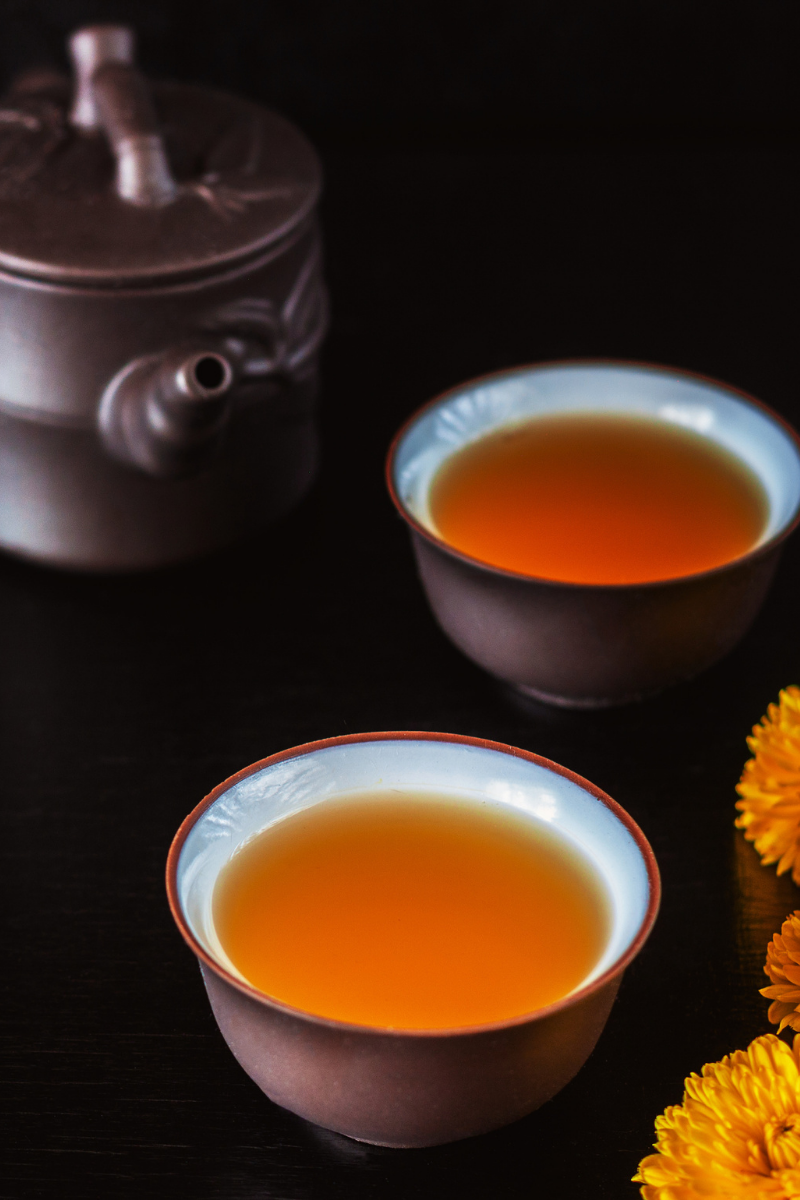 Two cups of amber-colored tea, a gray teapot, and yellow flowers on a dark surface.