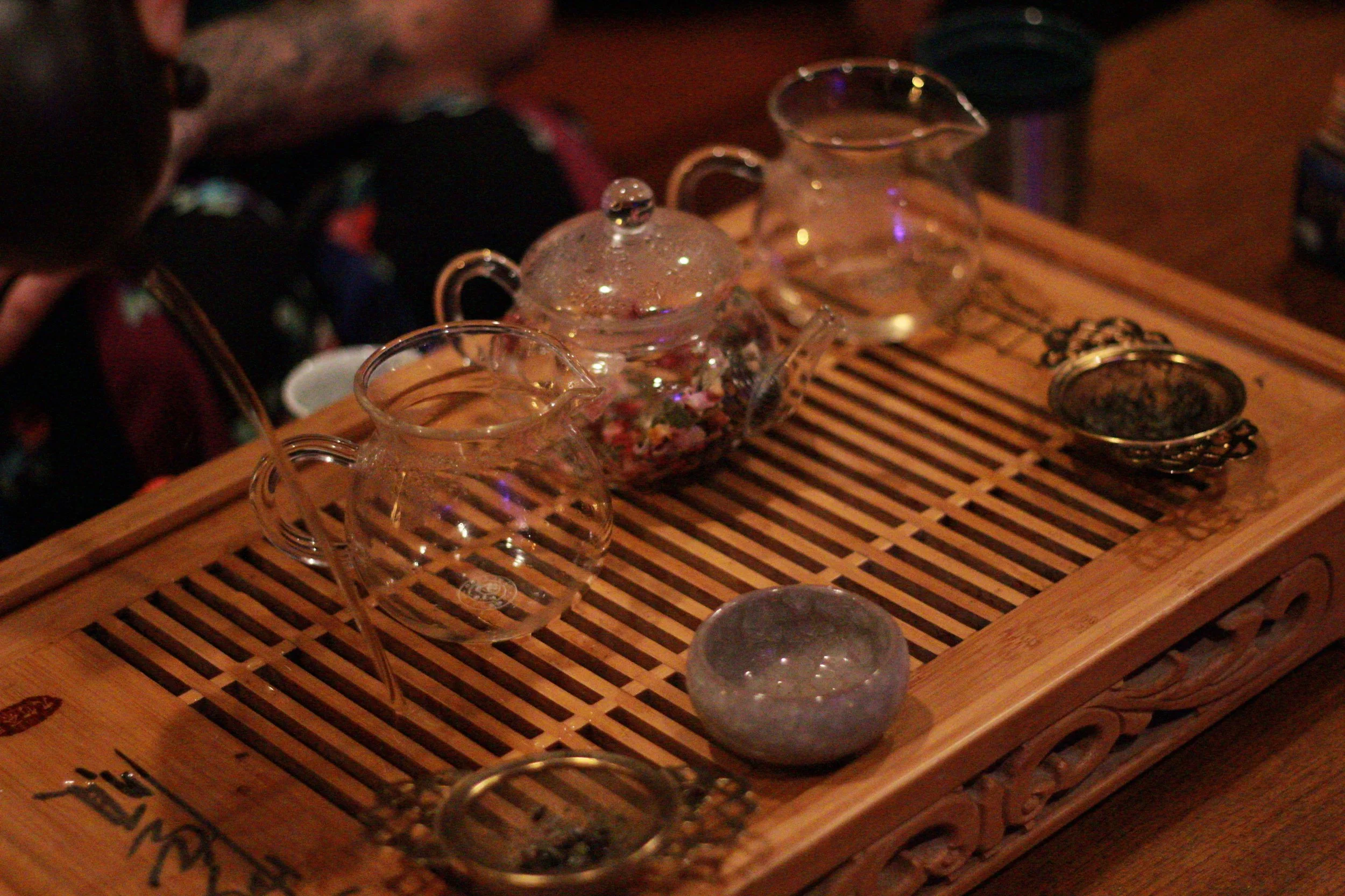 Glass teapot and cups on a wooden tray, with a person partially visible on the left.