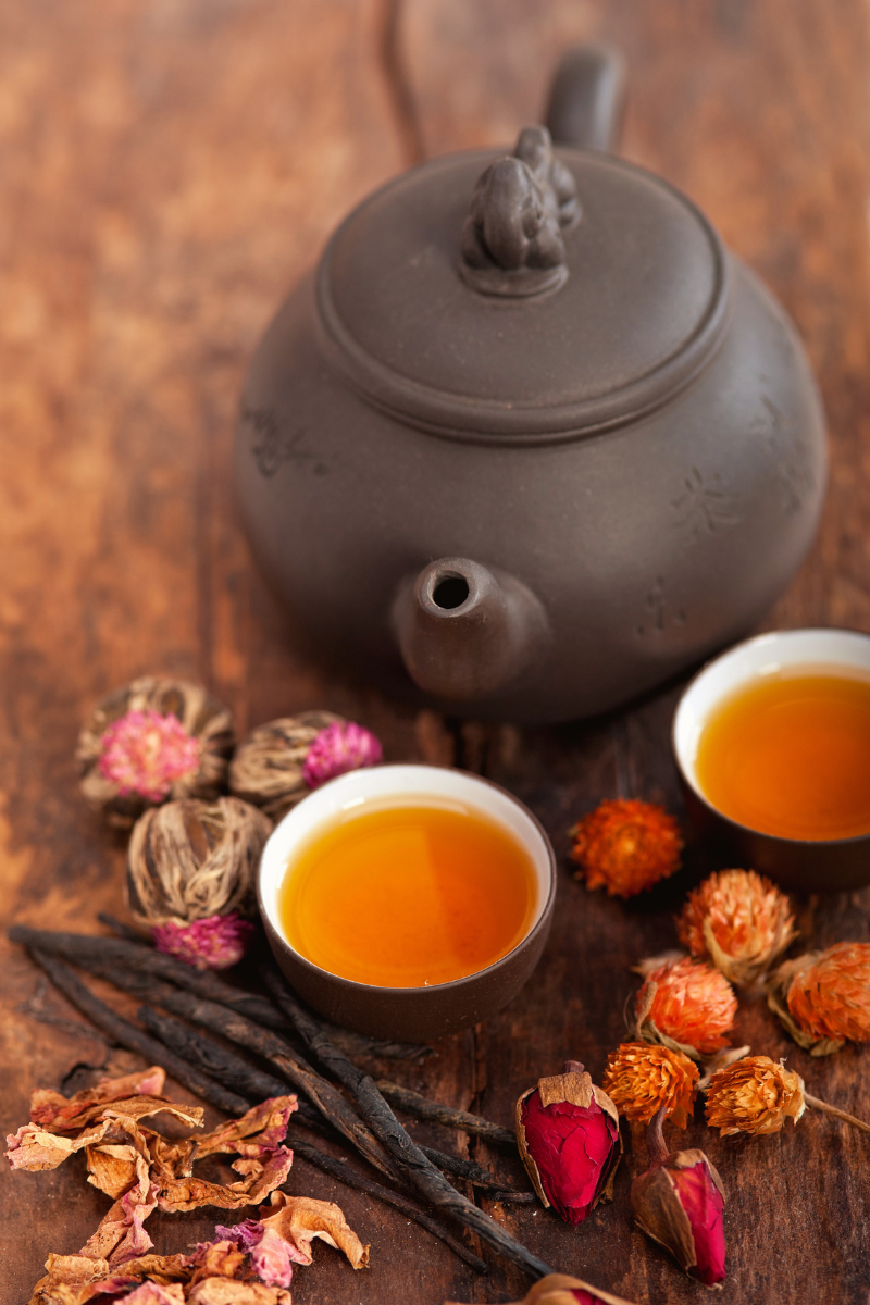 A black teapot with a small handle, two cups of amber-colored tea, and assorted dried flowers and herbs on a wooden surface.