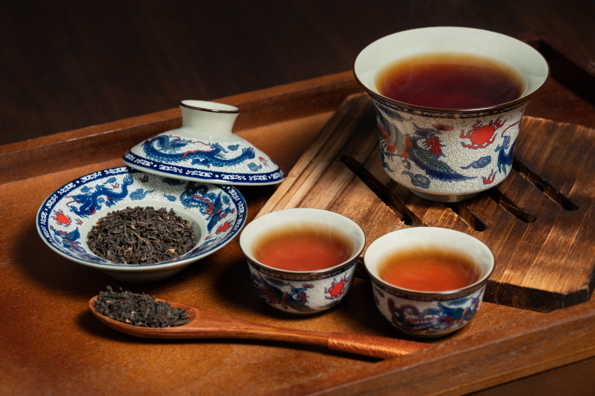A traditional Chinese tea set with a teacup filled with dark tea, two smaller teacups with tea, a bowl of loose black tea leaves, and a wooden spoon with loose tea, all arranged on a wooden tray.