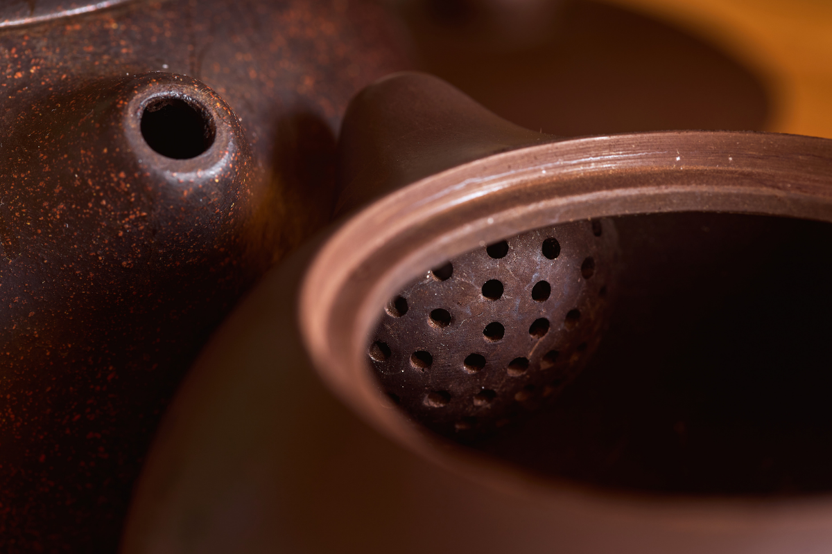 Close-up of a rusty, dark-colored ceramic teapot, showing the spout and the lid's underside with small holes.