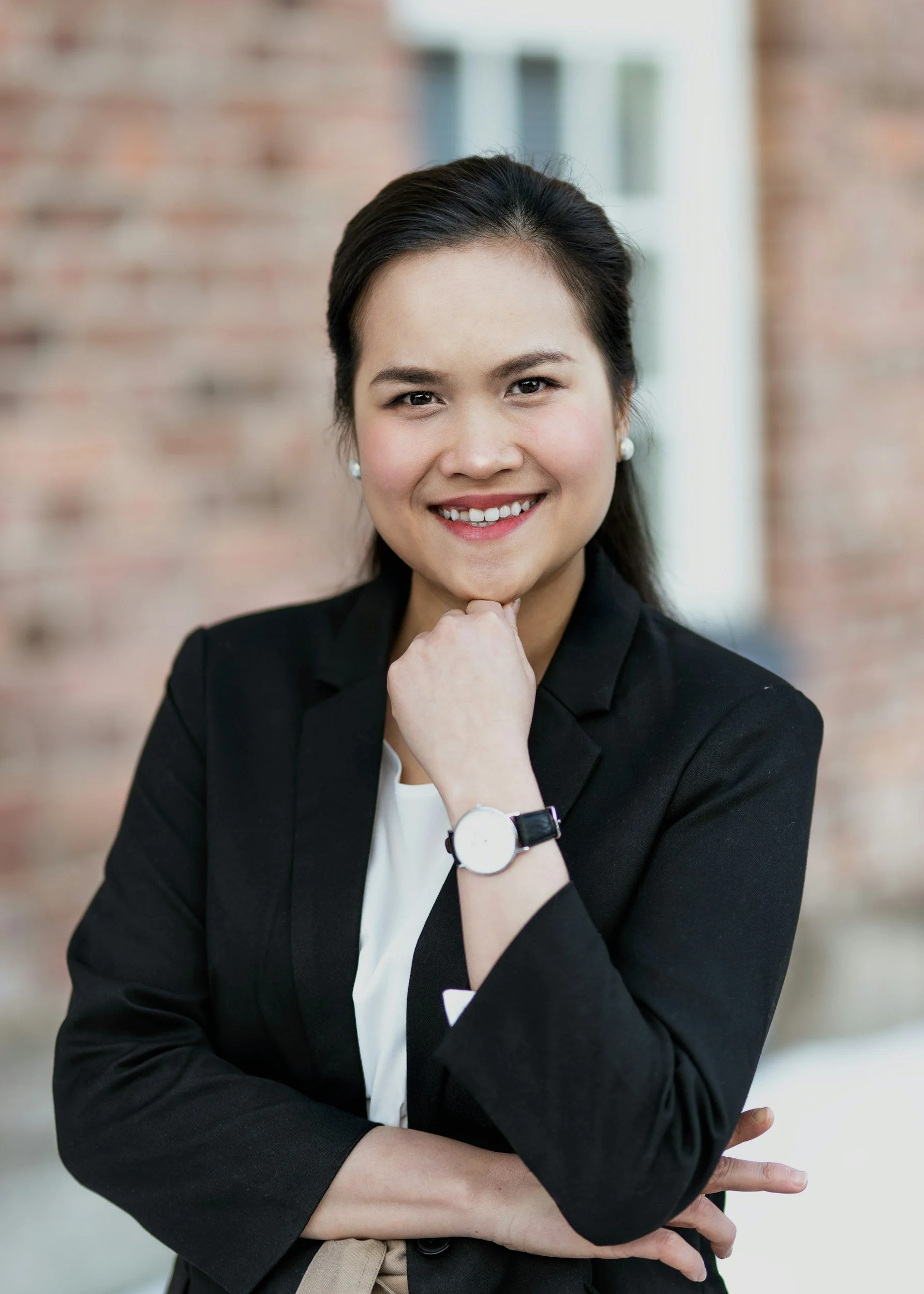 A woman with dark hair styled back, wearing a black blazer, white shirt, pearl earrings, and a white watch, smiling and posing with her hand under her chin in front of a blurred brick wall background.