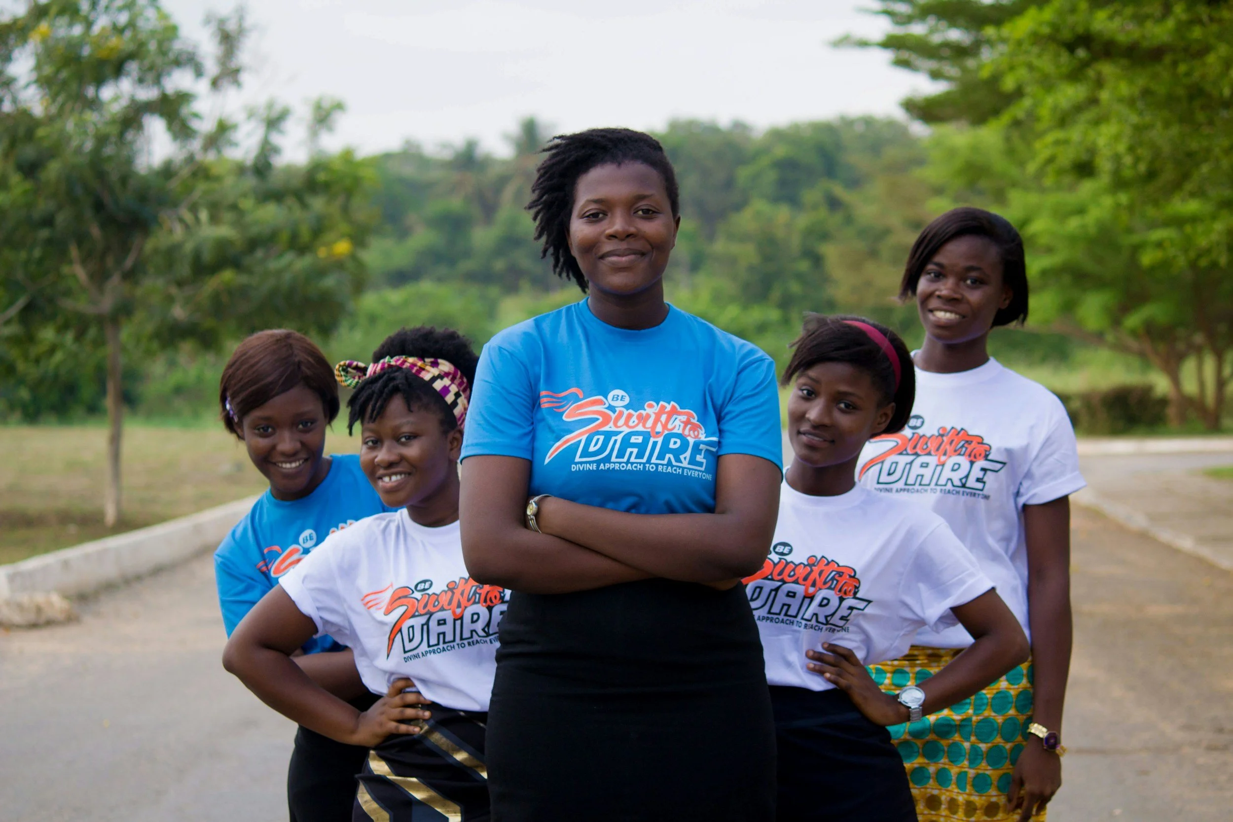 A group of five smiling young Black women outdoors, wearing matching white and blue t-shirts with the text 'Swift to Dare' in orange letters, standing with arms folded or at their sides, with lush green trees in the background.