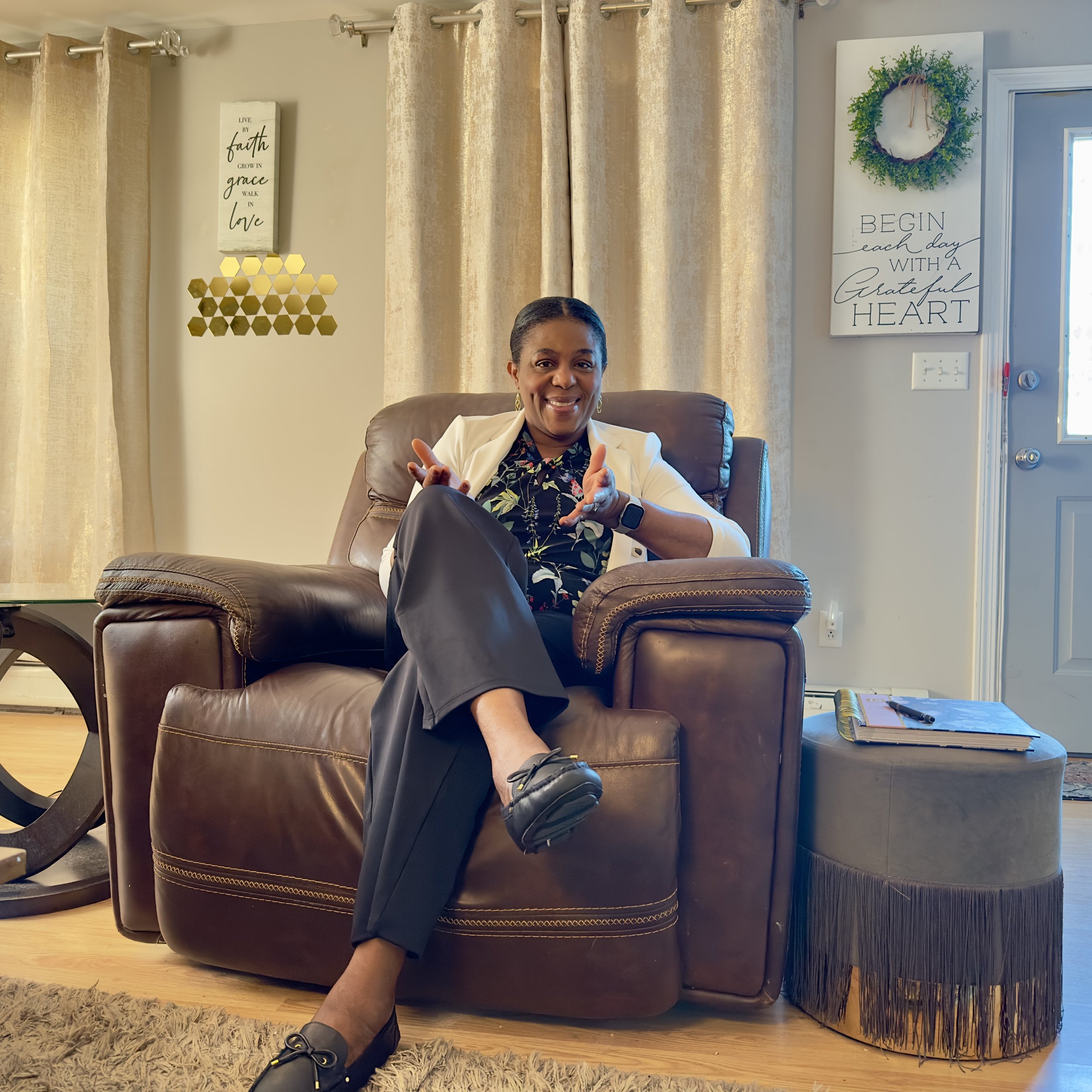A woman sitting on a brown leather couch, smiling, with her hands gesturing towards the camera. She is wearing a white blazer over a floral blouse, black pants, and black shoes. The room has beige curtains, wall decorations with inspirational quotes, a beige shag rug, and a side table with a book and pen.