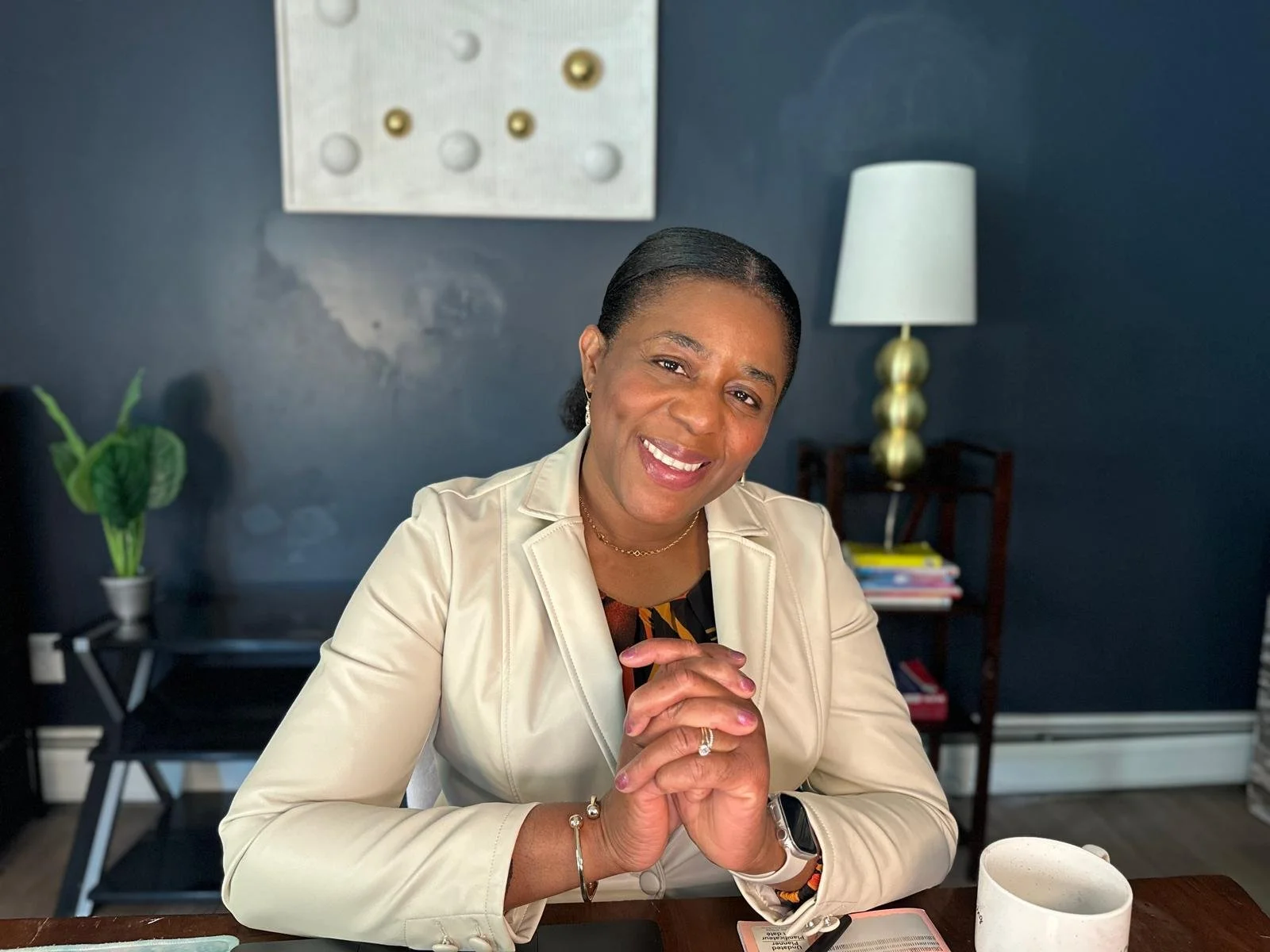 A woman with dark hair pulled back, wearing a beige blazer, sitting at a wooden table and smiling at the camera, with a dark blue wall, a white and gold lamp, a small shelf with books, and a potted plant in the background.