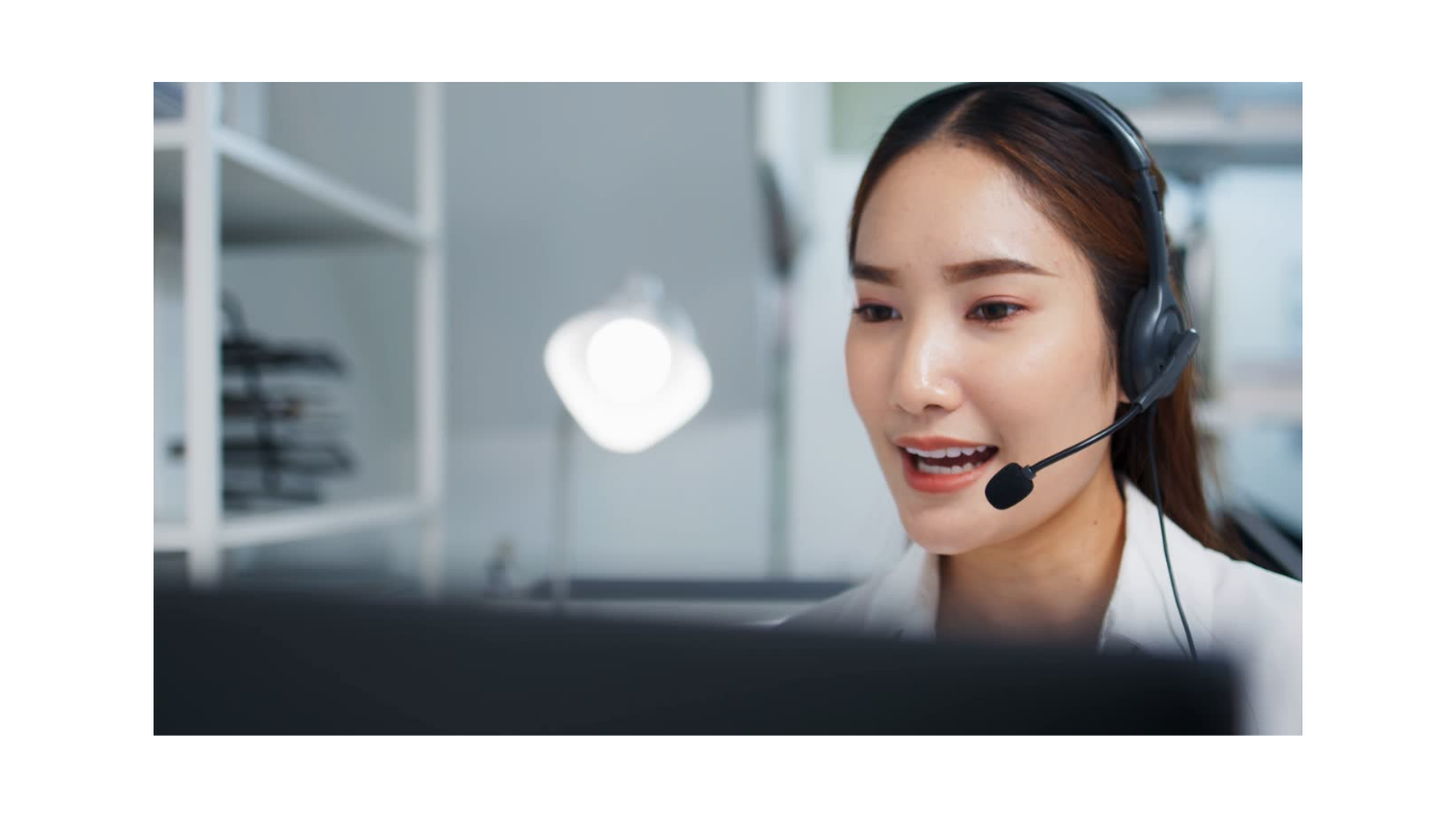 A woman wearing a headset with a microphone, smiling and looking at her computer screen in an office environment.