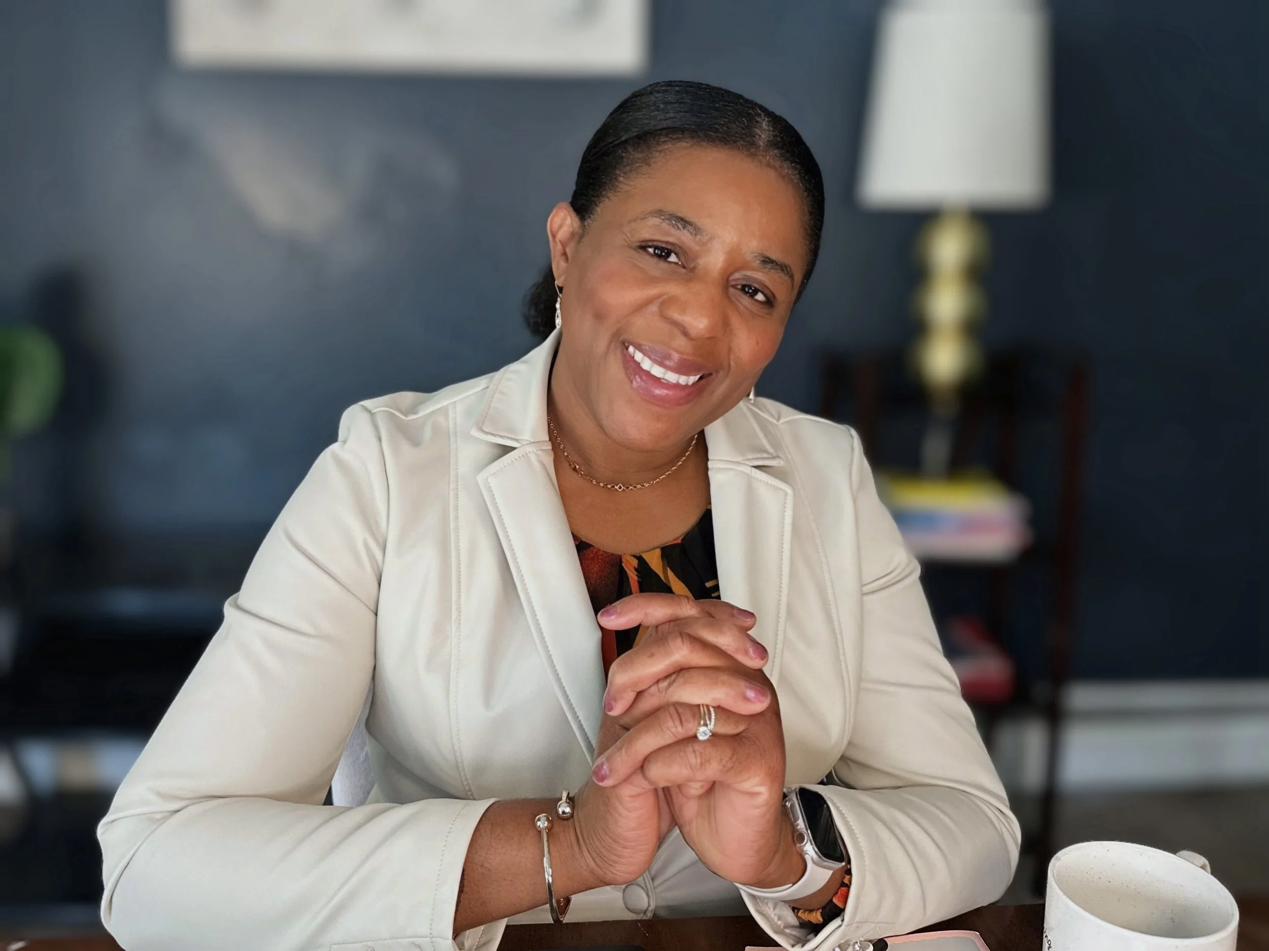 A smiling woman with dark hair slicked back, wearing a white blazer, sitting at a table with hands clasped. In the background, there is a lamp and a dark blue wall.