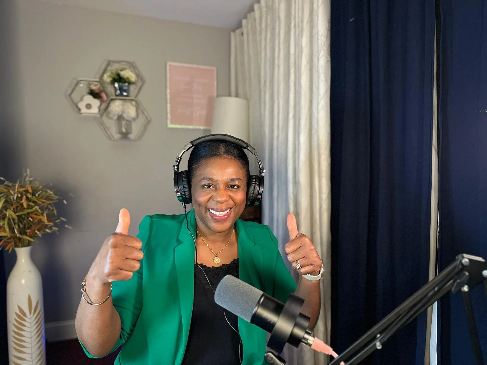 A woman wearing headphones and a green blazer giving a double thumbs up in a recording studio.