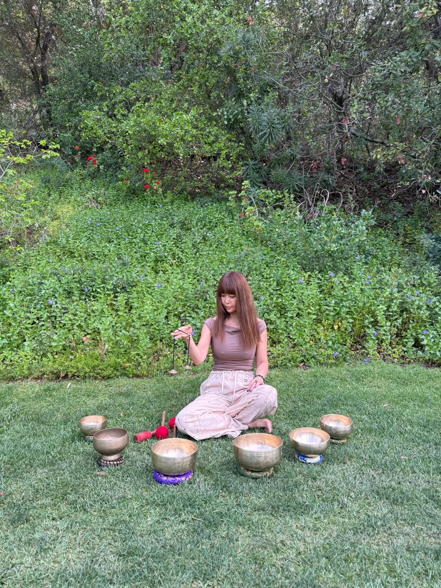A woman sitting on grass in a garden, holding a singing bowl, surrounded by other bowls and mallets, with a backdrop of dense green bushes and trees.