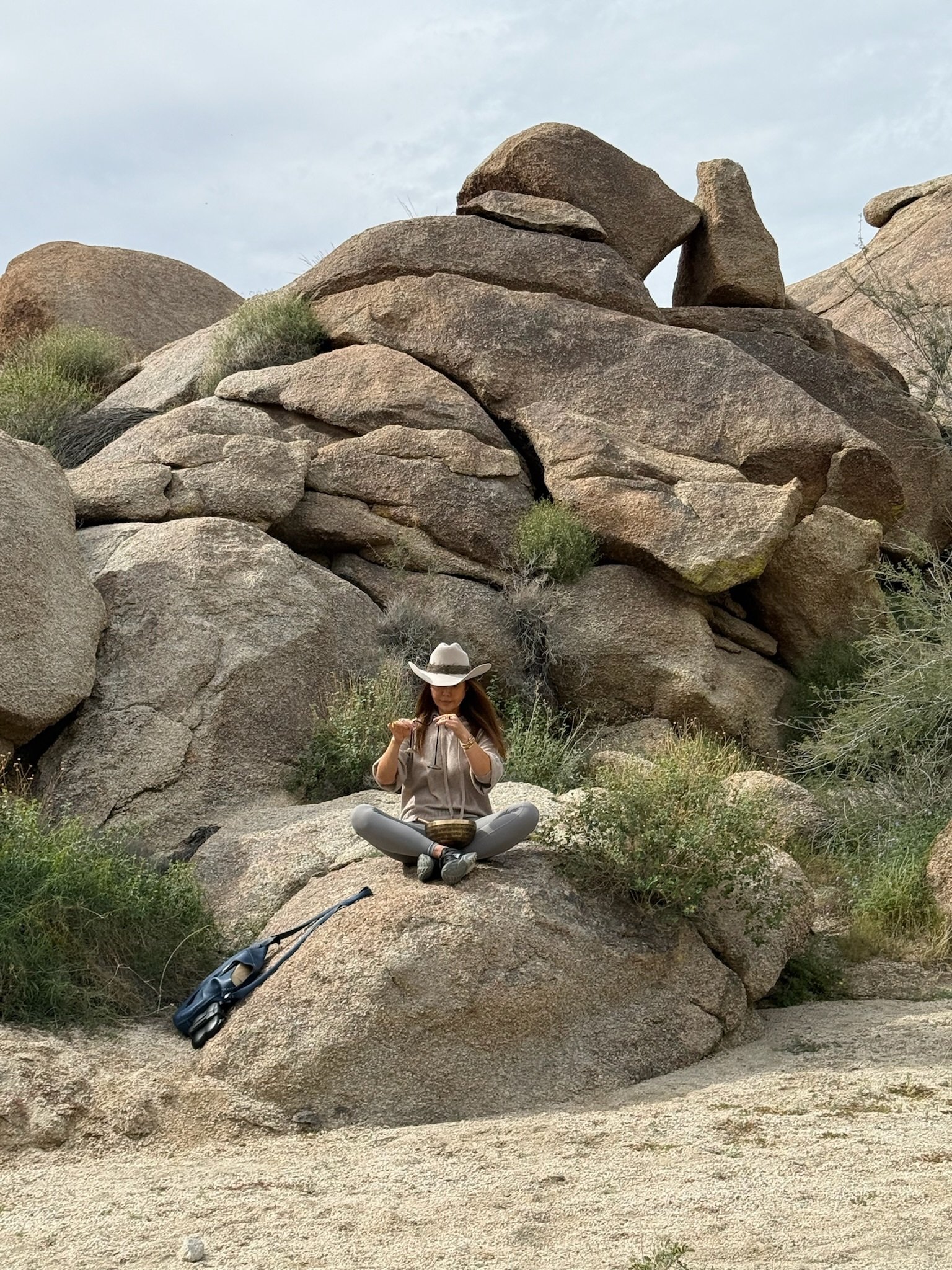 A woman sitting on a large rock at a desert with large boulders and bushes, wearing a wide-brimmed hat, gray pants, a light shirt, and holding a bowl and a spoon.