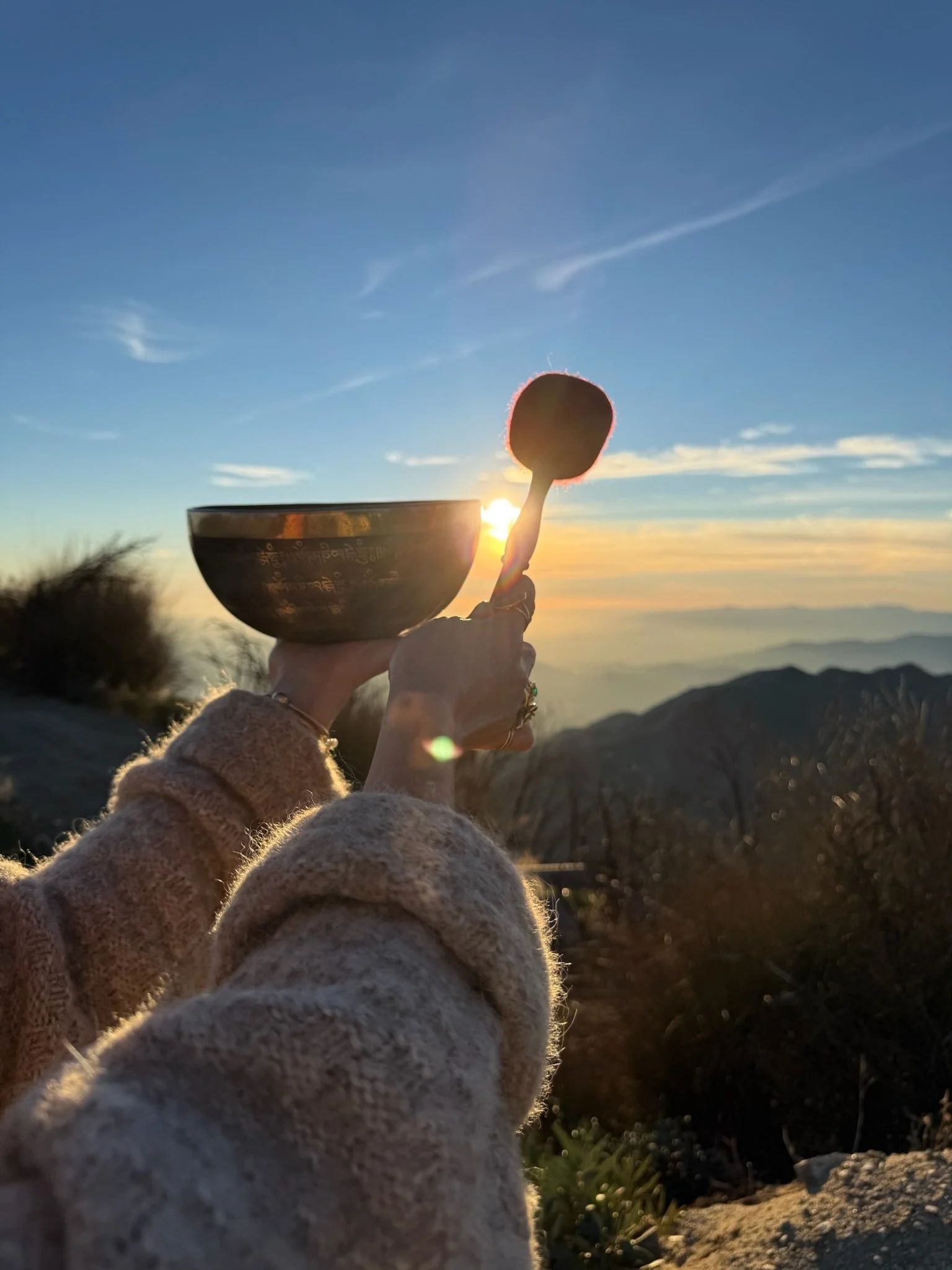 Person holding a Tibetan singing bowl and a mallet during sunset with mountains in the background.