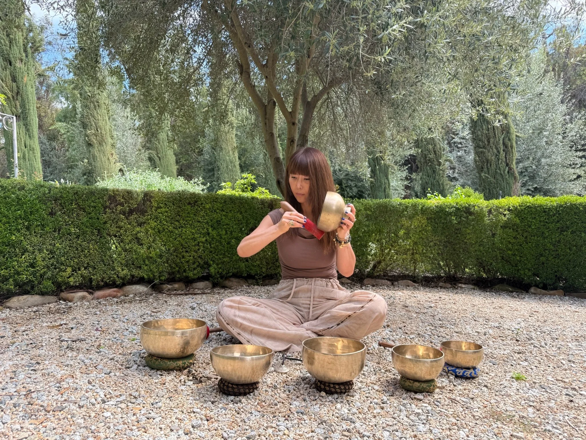 A woman sitting cross-legged outdoors on gravel, playing singing bowls surrounded by greenery and trees.